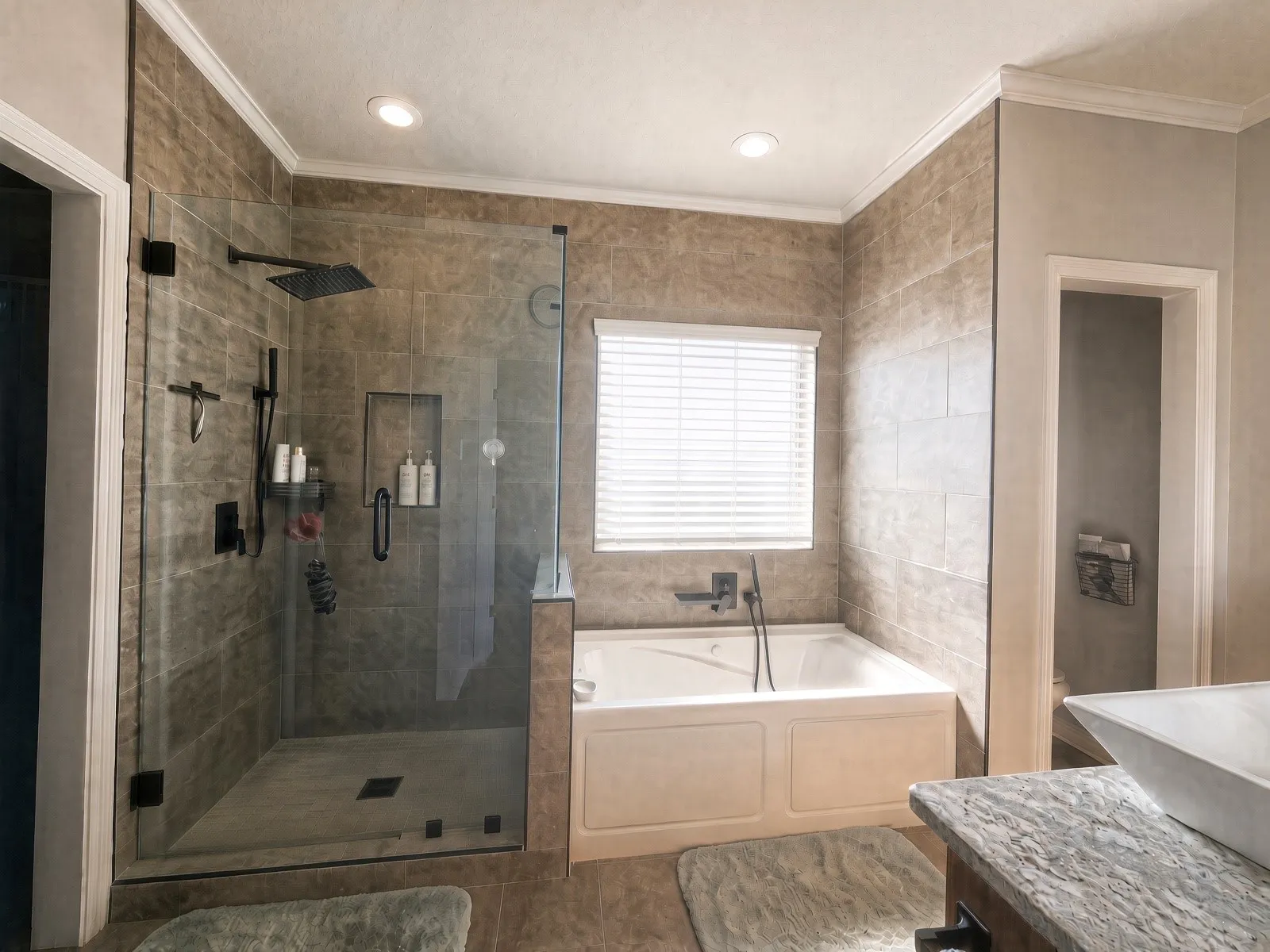 Walk-in shower with clear glass enclosure and black hardware, soaking tub with integrated faucet, recessed ceiling lighting, crown molding, and tiled walls