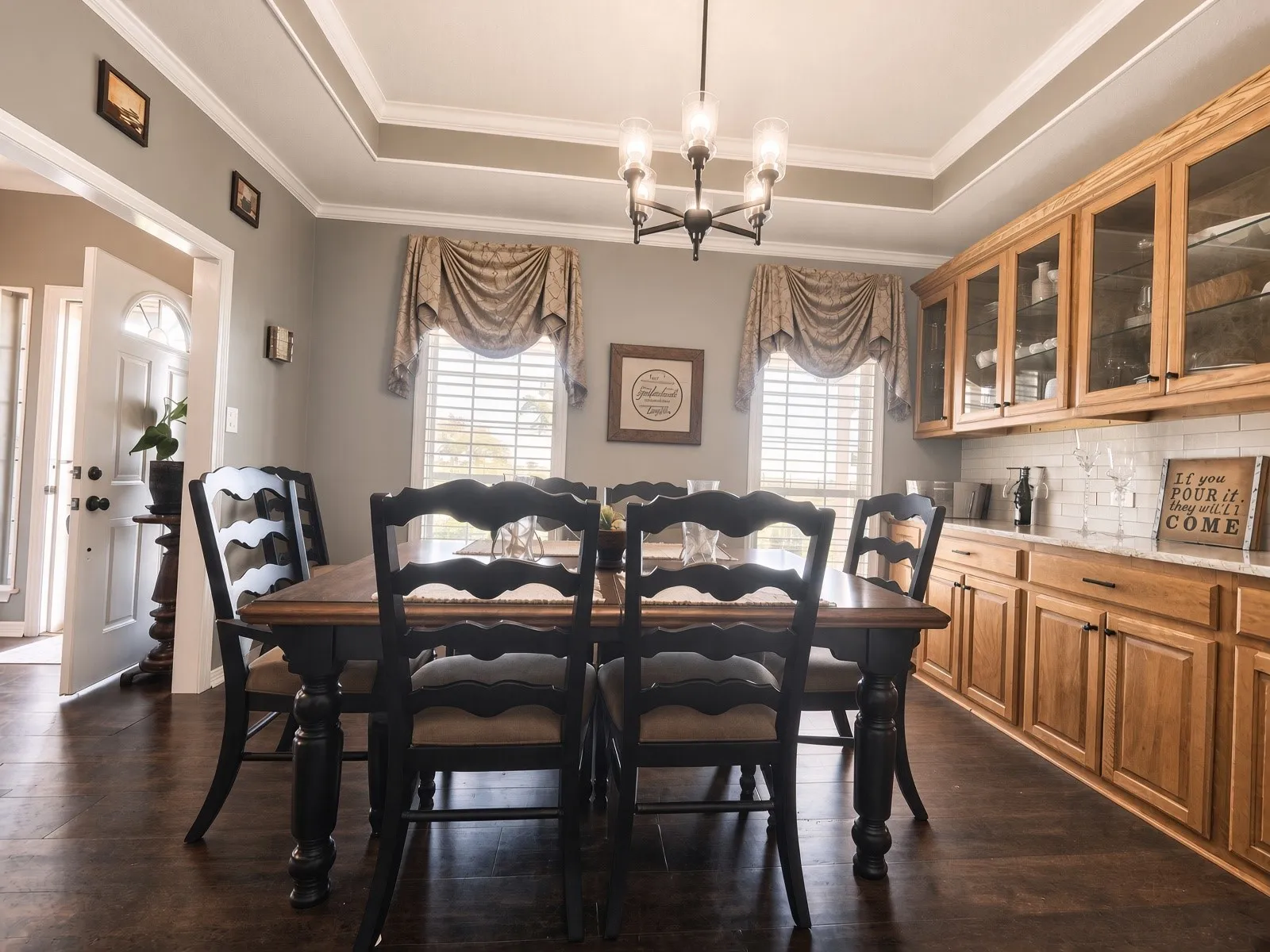 Tray ceiling with crown molding, dark wood-finish flooring, built-in cabinetry with glass-front uppers, subway tile backsplash, and a contemporary chandelier