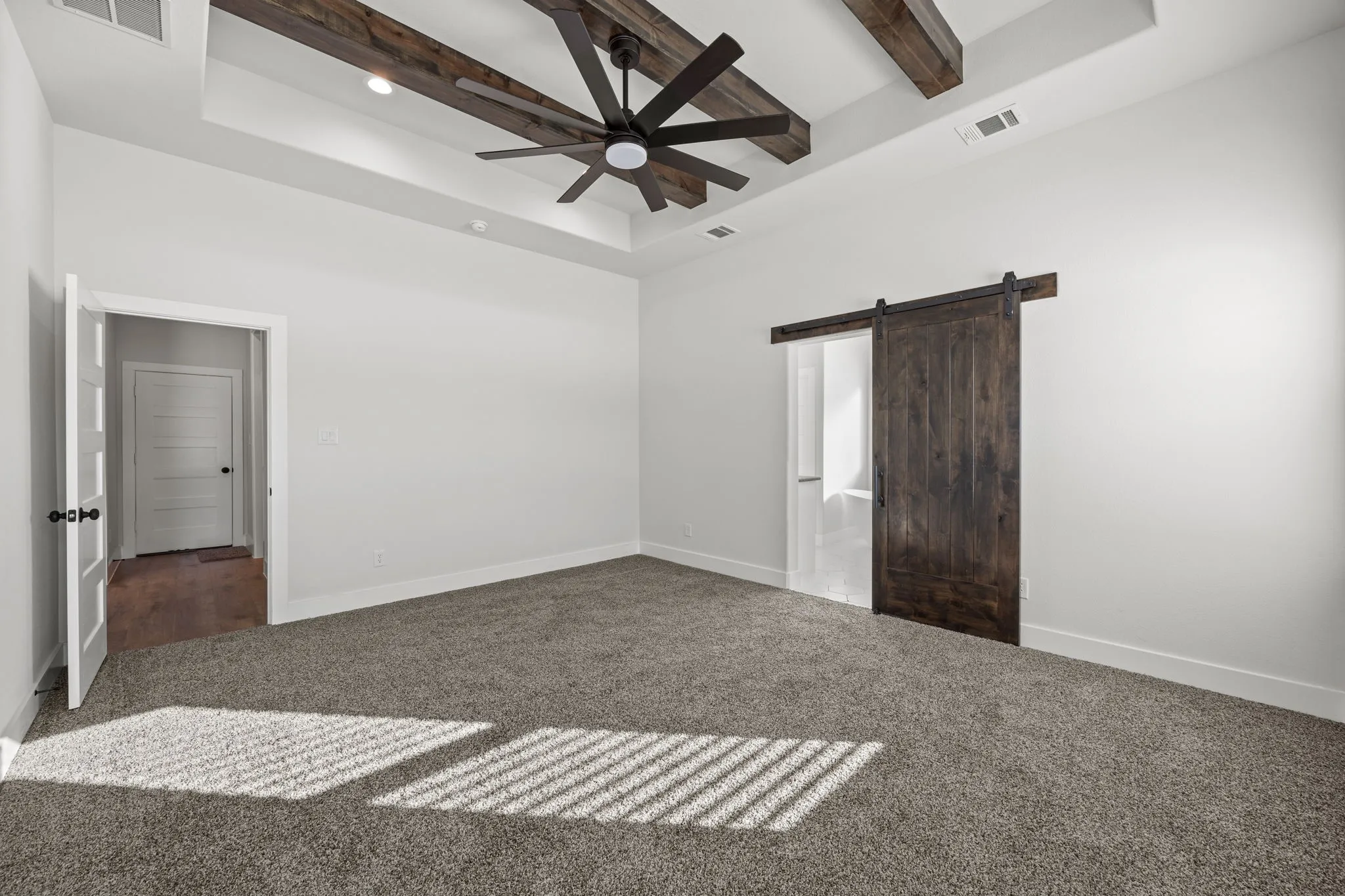 Unfurnished bedroom with a barn door, a tray ceiling, carpet flooring, a ceiling fan, and beam ceiling