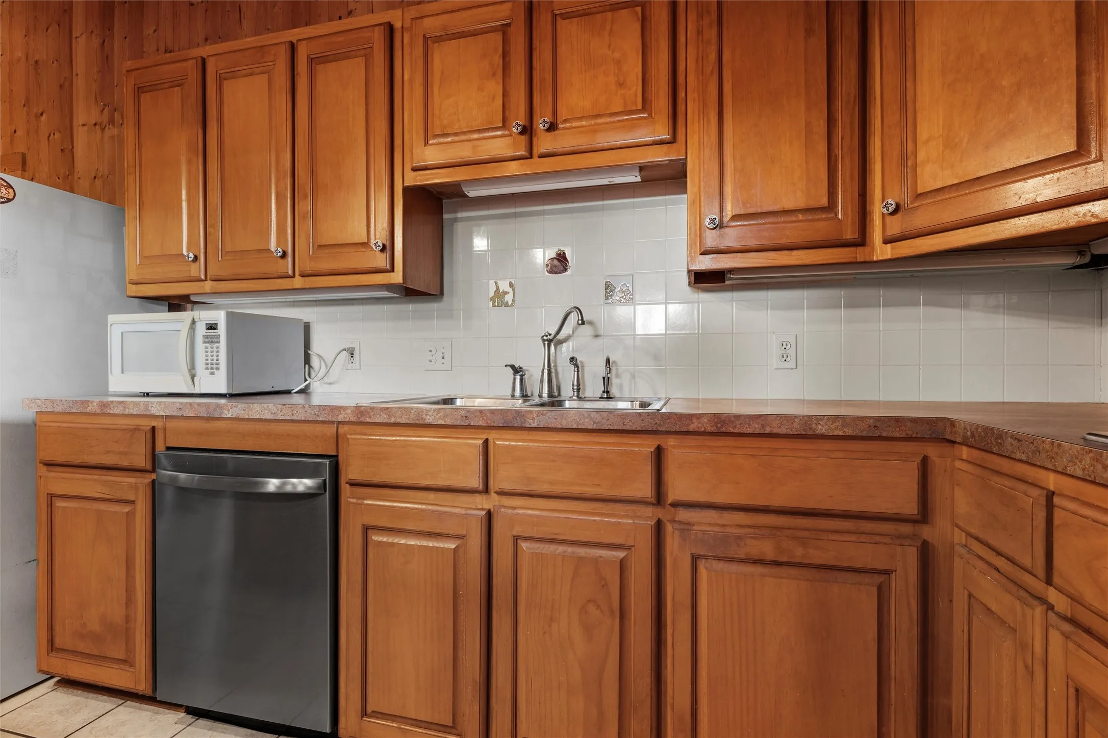 Kitchen featuring brown cabinetry, backsplash, dishwasher, white microwave, and light tile patterned floors