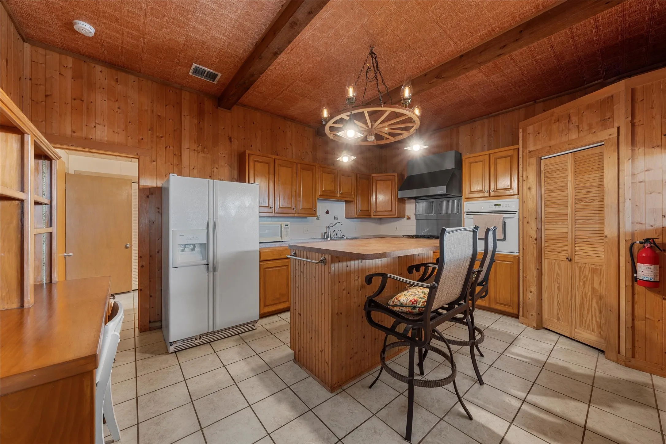 Kitchen with wood walls, white appliances, wall chimney exhaust hood, light tile patterned flooring, and a breakfast bar