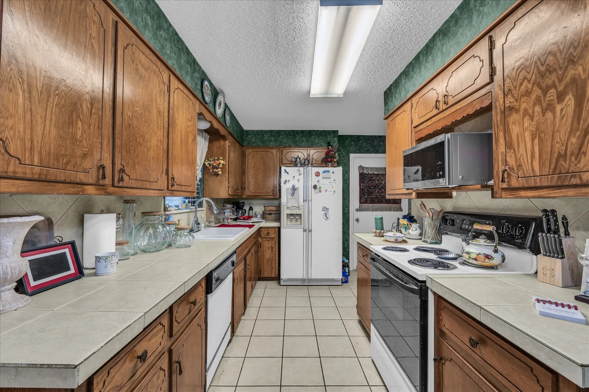 Kitchen featuring white appliances, brown cabinetry, light tile patterned flooring, a textured ceiling, and backsplash