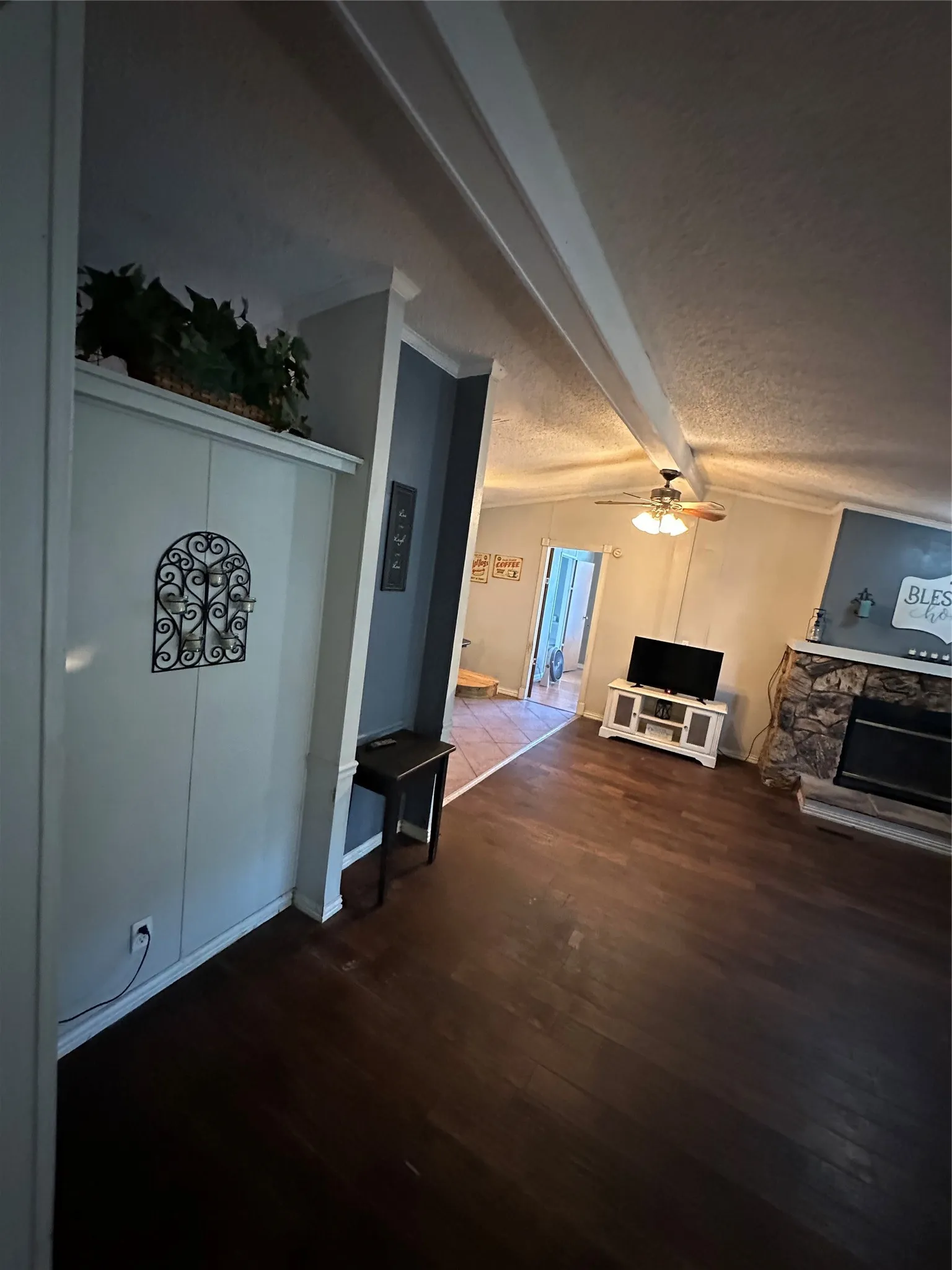 Living area with beam ceiling, a textured ceiling, dark wood-style floors, a ceiling fan, and a stone fireplace