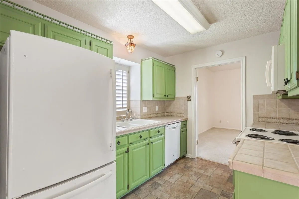 Kitchen with green cabinets, white appliances, tile counters, light stone finish floors, and backsplash