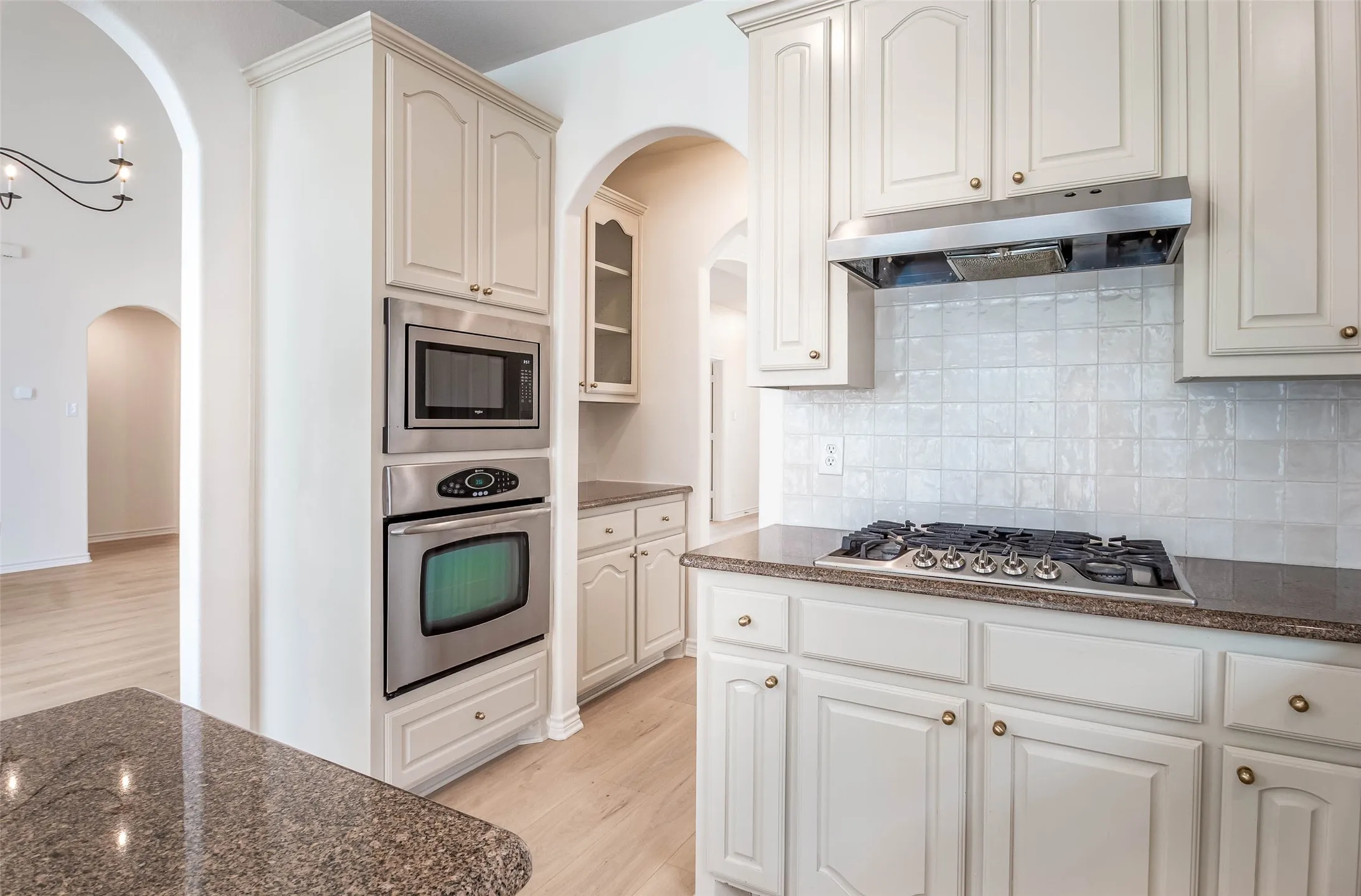 Kitchen with light wood-style floors, arched walkways, appliances with stainless steel finishes, tasteful backsplash, and under cabinet range hood