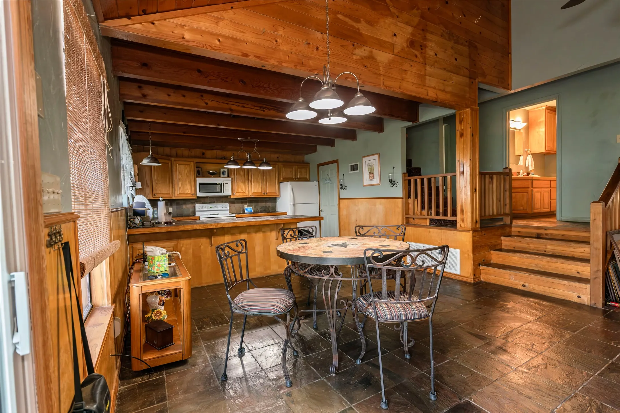 Kitchen featuring brown cabinetry, hanging light fixtures, wainscoting, beamed ceiling, and white appliances