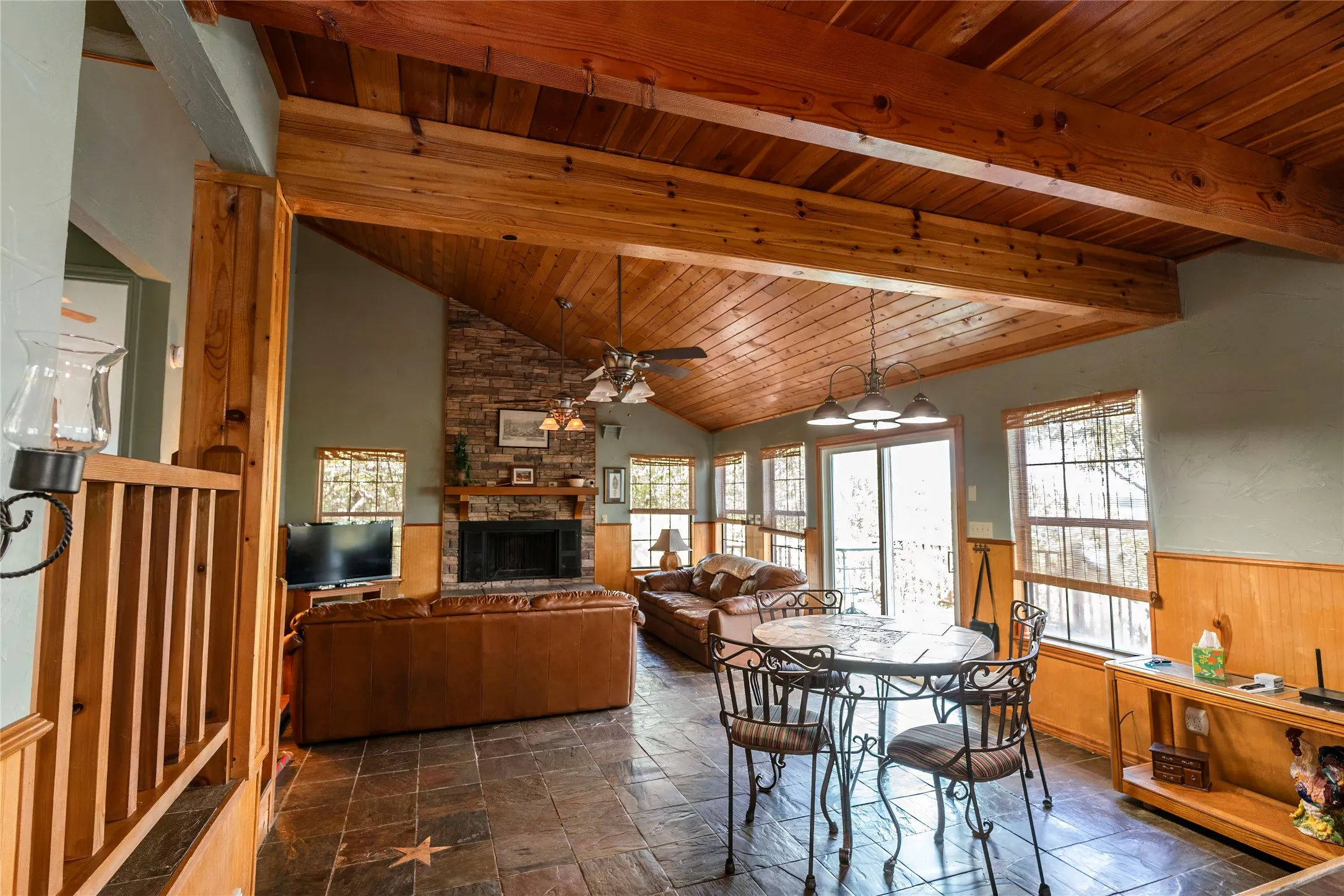 Dining room with a stone fireplace, a wood ceiling with exposed beams, a ceiling fan, stone tile flooring, and a wainscoted wall
