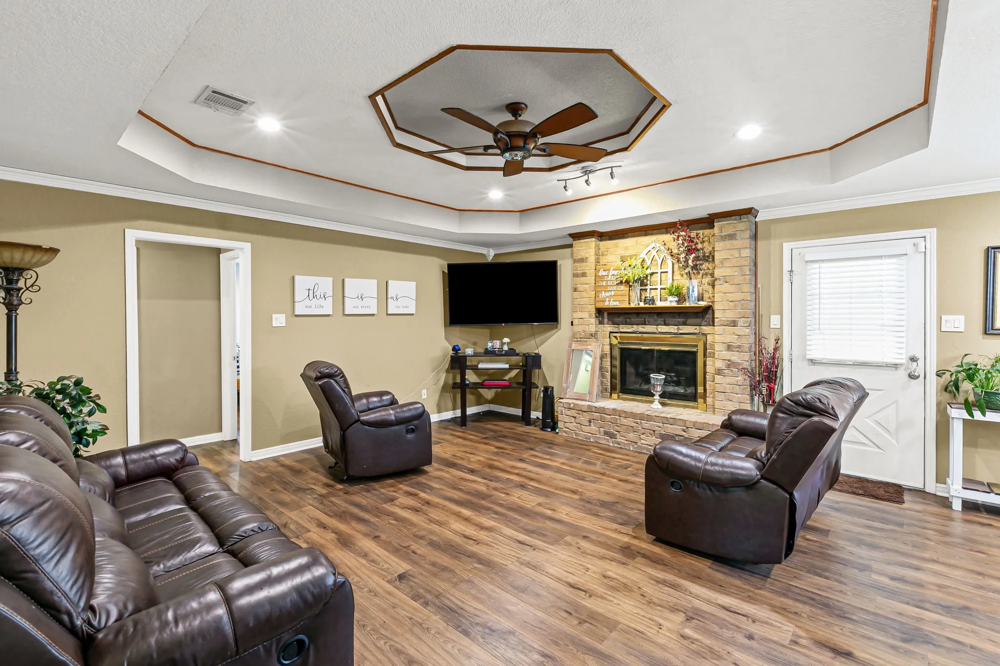 Living area featuring a raised ceiling, a brick fireplace, dark wood-style floors, a ceiling fan, and crown molding