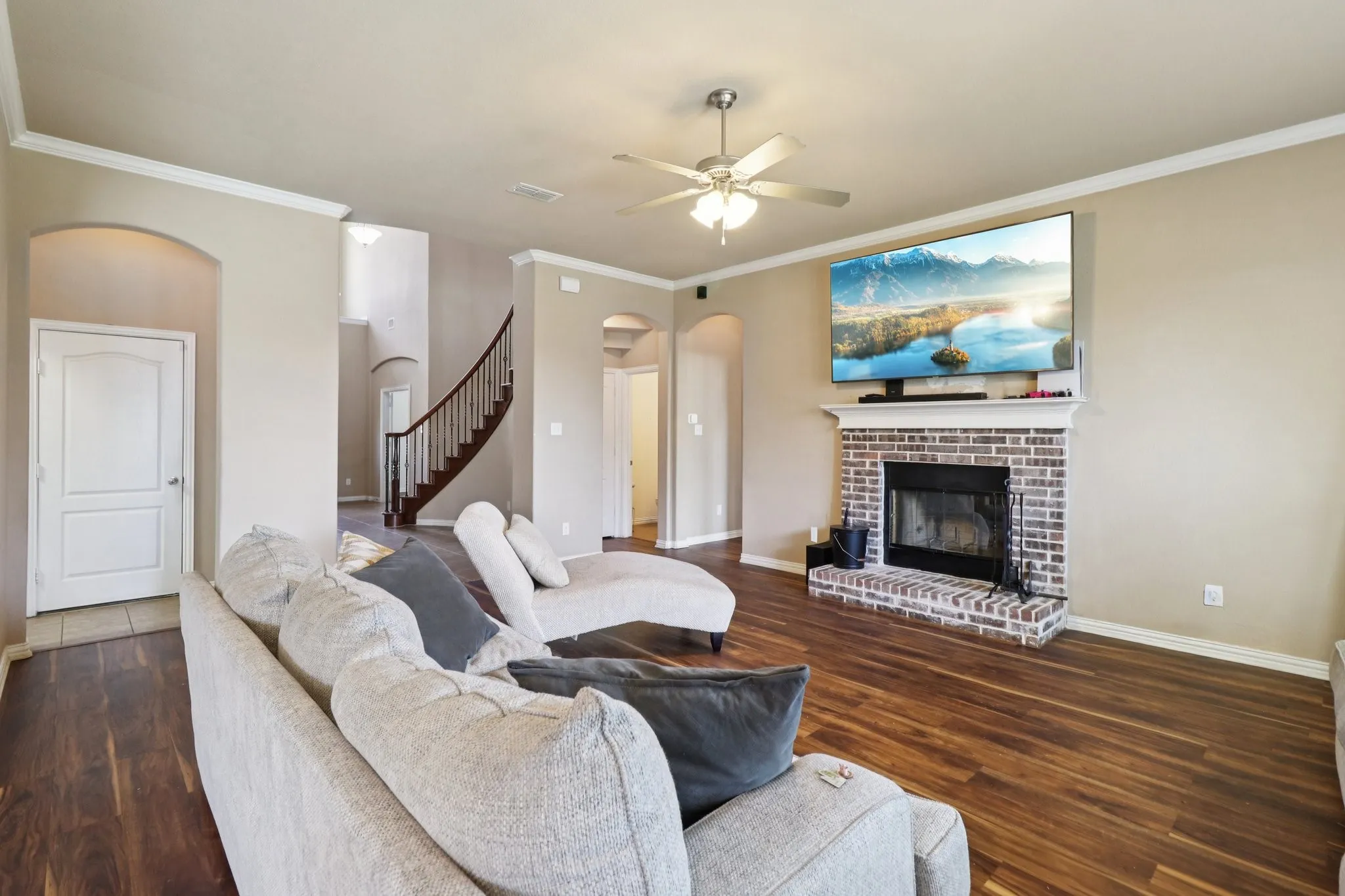Living room featuring crown molding, a brick fireplace, stairway, a ceiling fan, and dark wood-style flooring