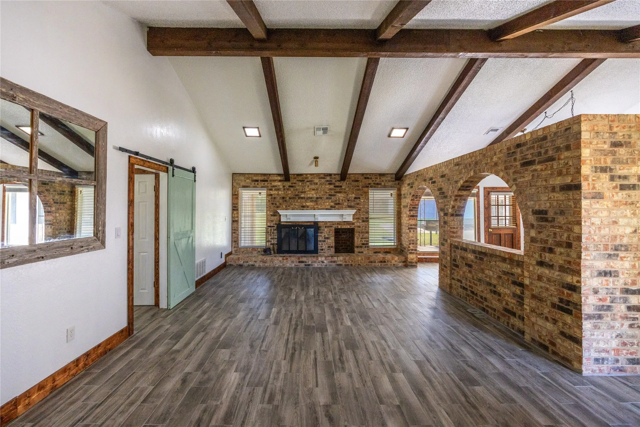 Unfurnished living room featuring a barn door, beam ceiling, dark wood-style flooring, an outdoor brick fireplace, and brick wall