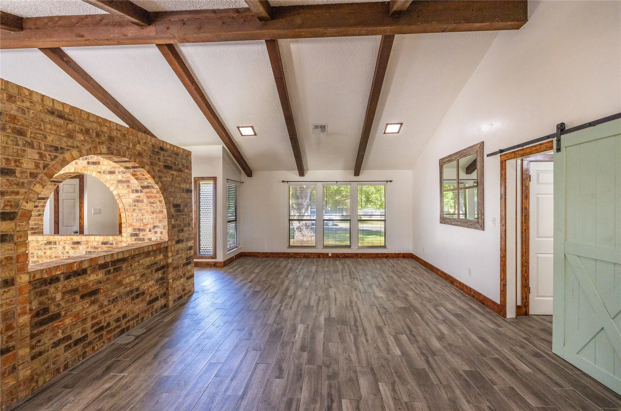 Unfurnished living room with a barn door, beam ceiling, dark wood-type flooring, brick wall, and high vaulted ceiling