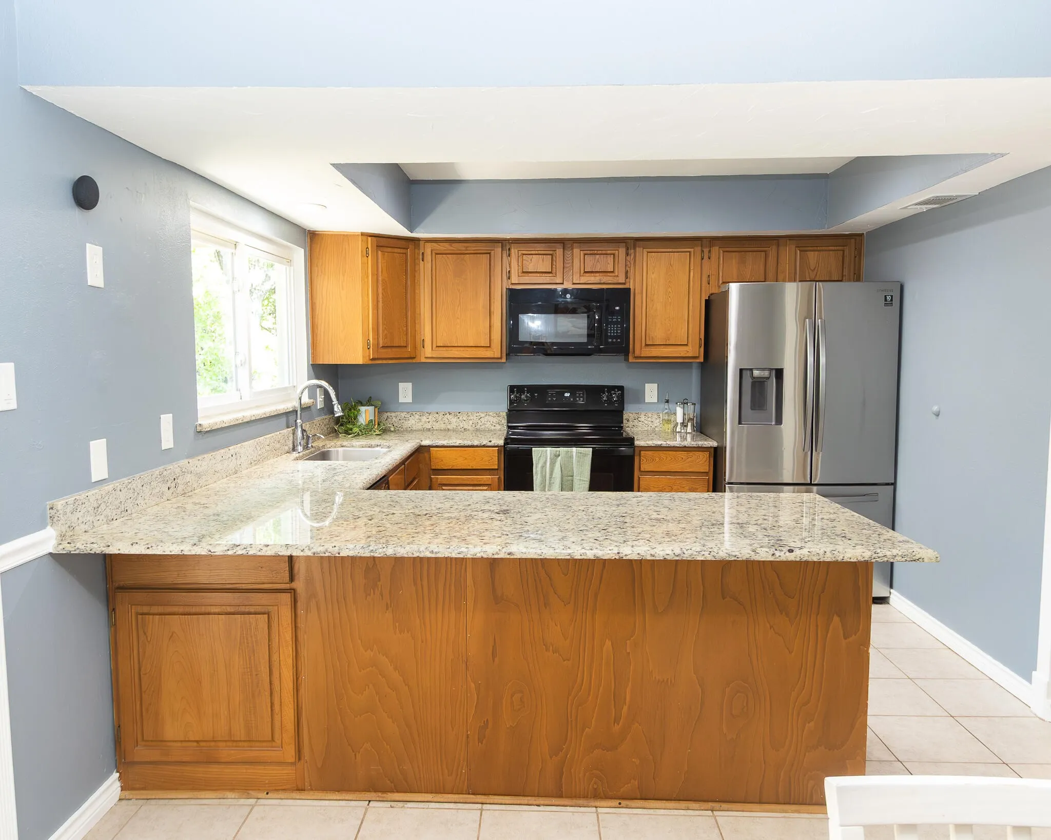 Kitchen featuring brown cabinetry, light stone counters, black appliances, light tile patterned floors, and a peninsula