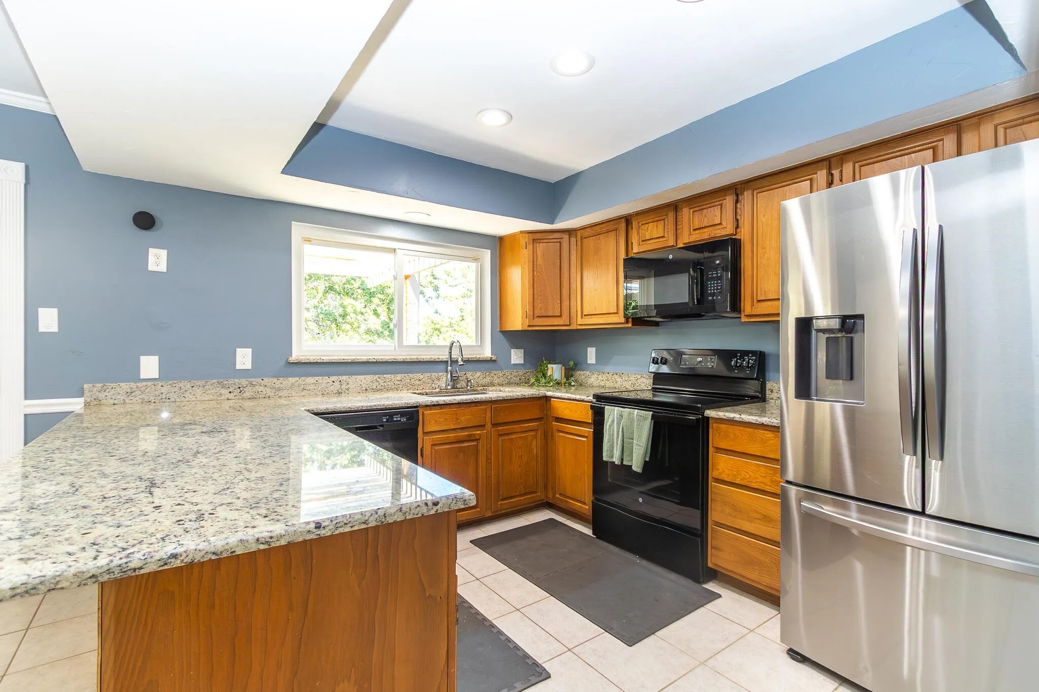 Kitchen with black appliances, brown cabinetry, light stone countertops, a peninsula, and light tile patterned flooring