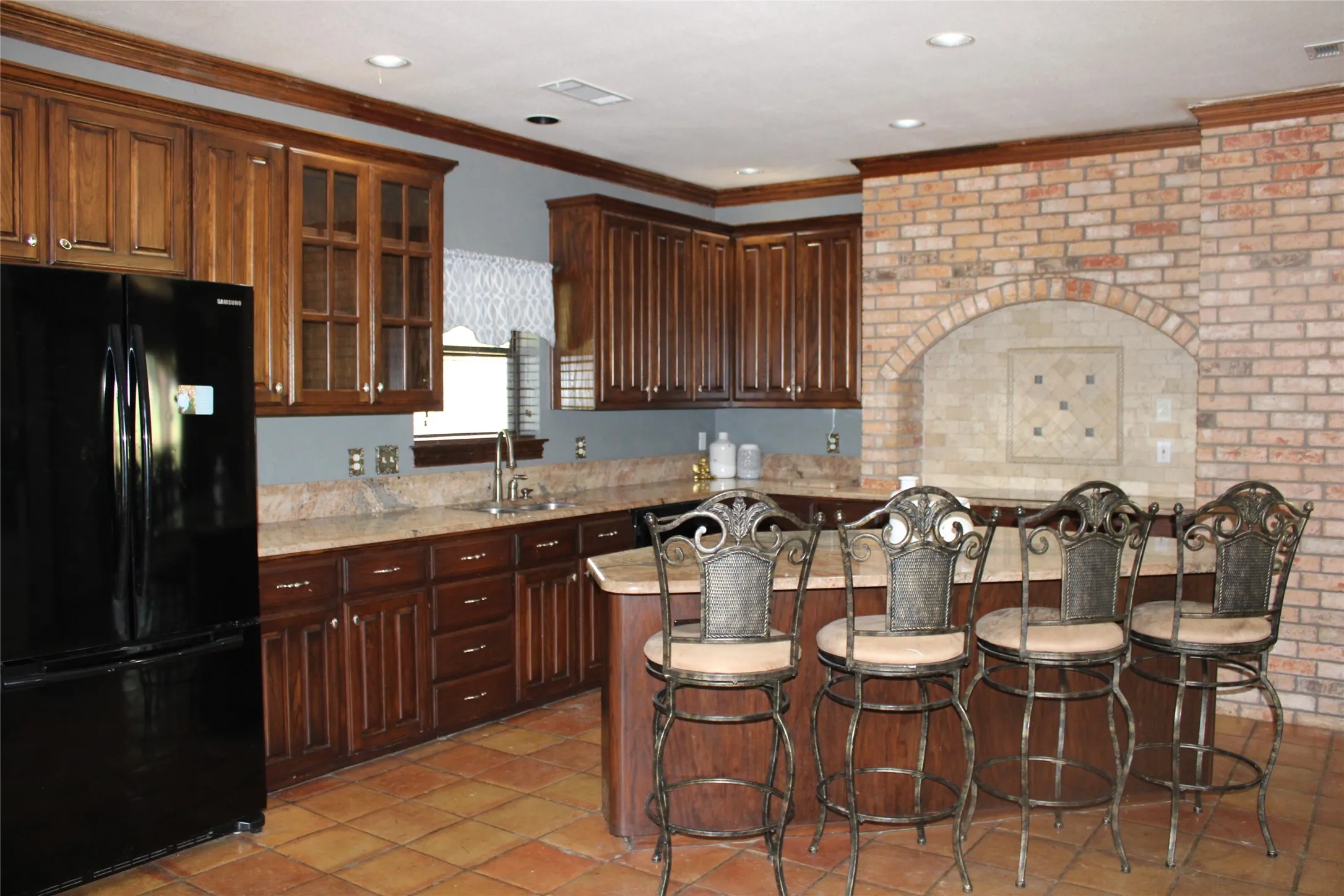 Kitchen with brick wall, black fridge, light stone countertops, light tile patterned floors, and sink