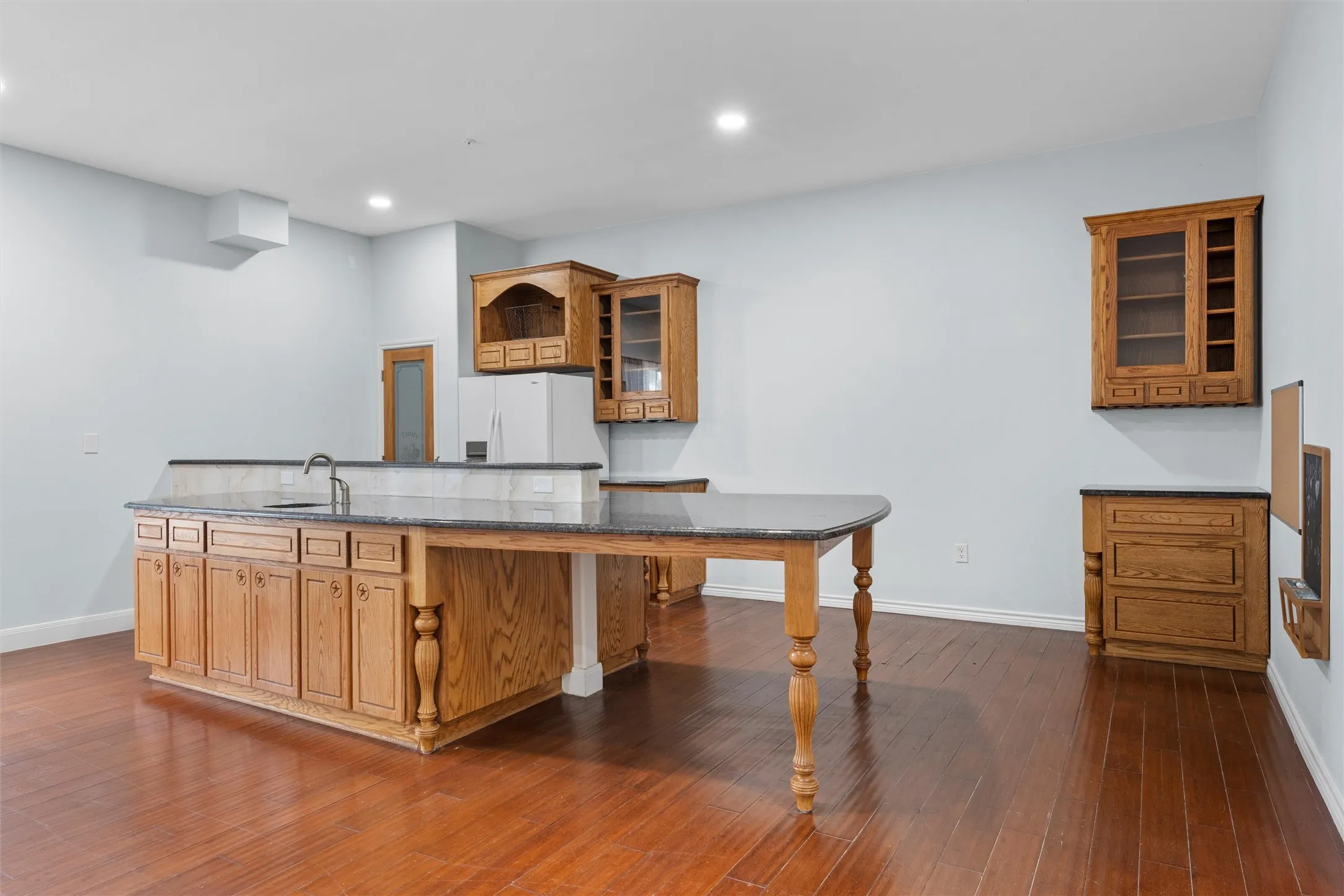 Kitchen featuring brown cabinets, glass insert cabinets, dark stone counters, dark wood-style flooring, and a kitchen island with sink