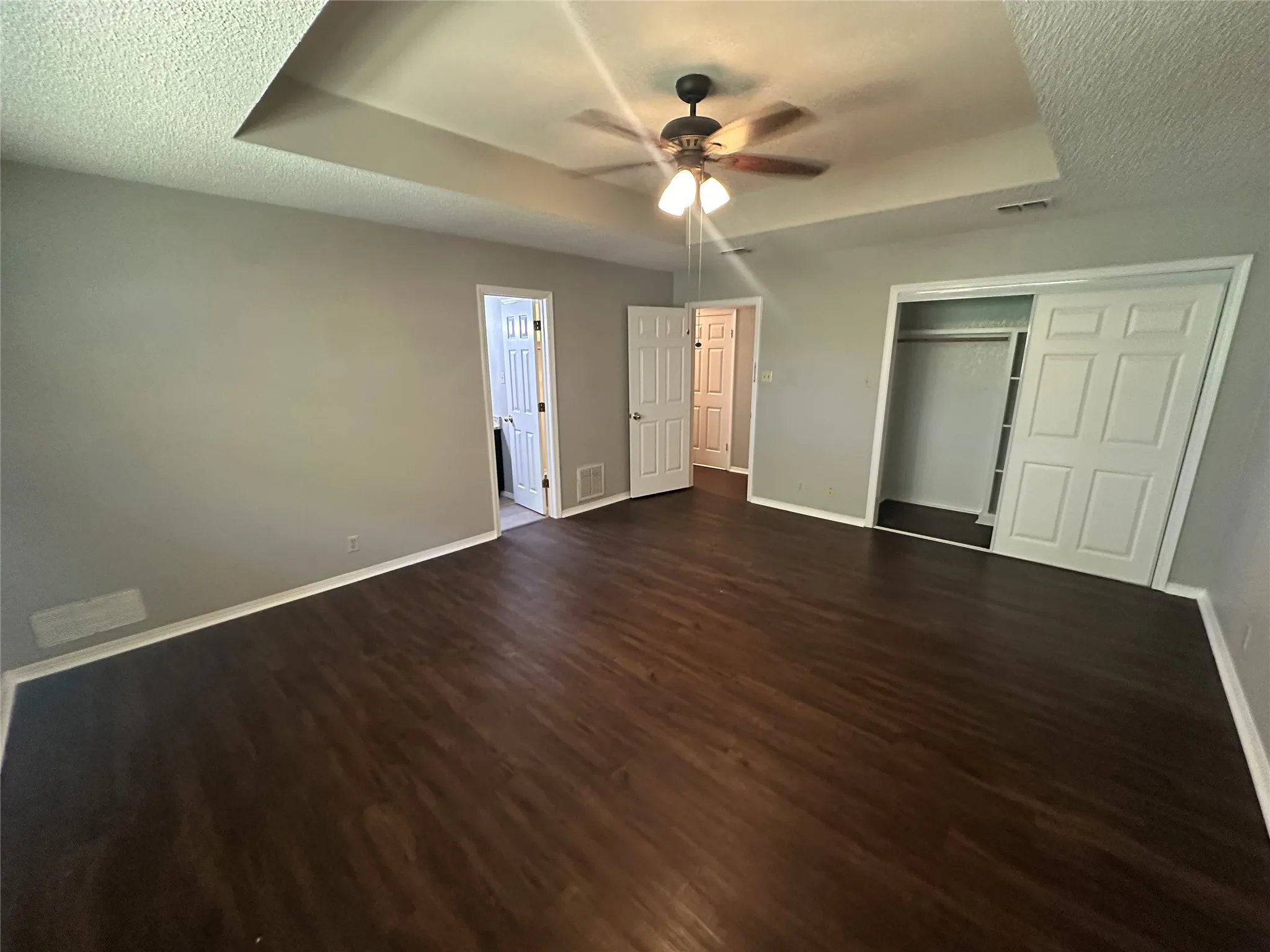 Unfurnished bedroom with a textured ceiling, a tray ceiling, dark wood-type flooring, a closet, and a ceiling fan