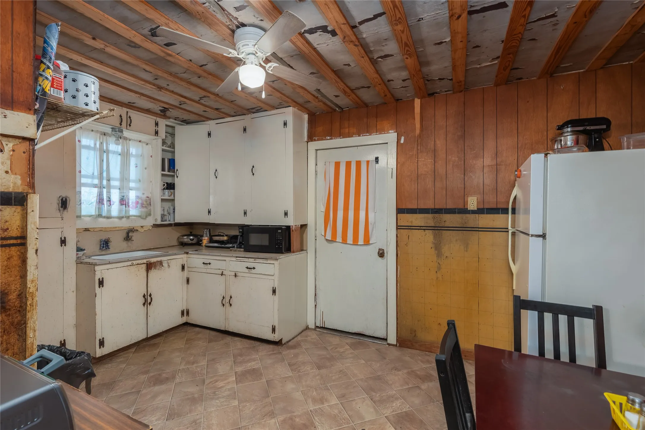 Kitchen with freestanding refrigerator, wooden walls, light countertops, black microwave, and ceiling fan
