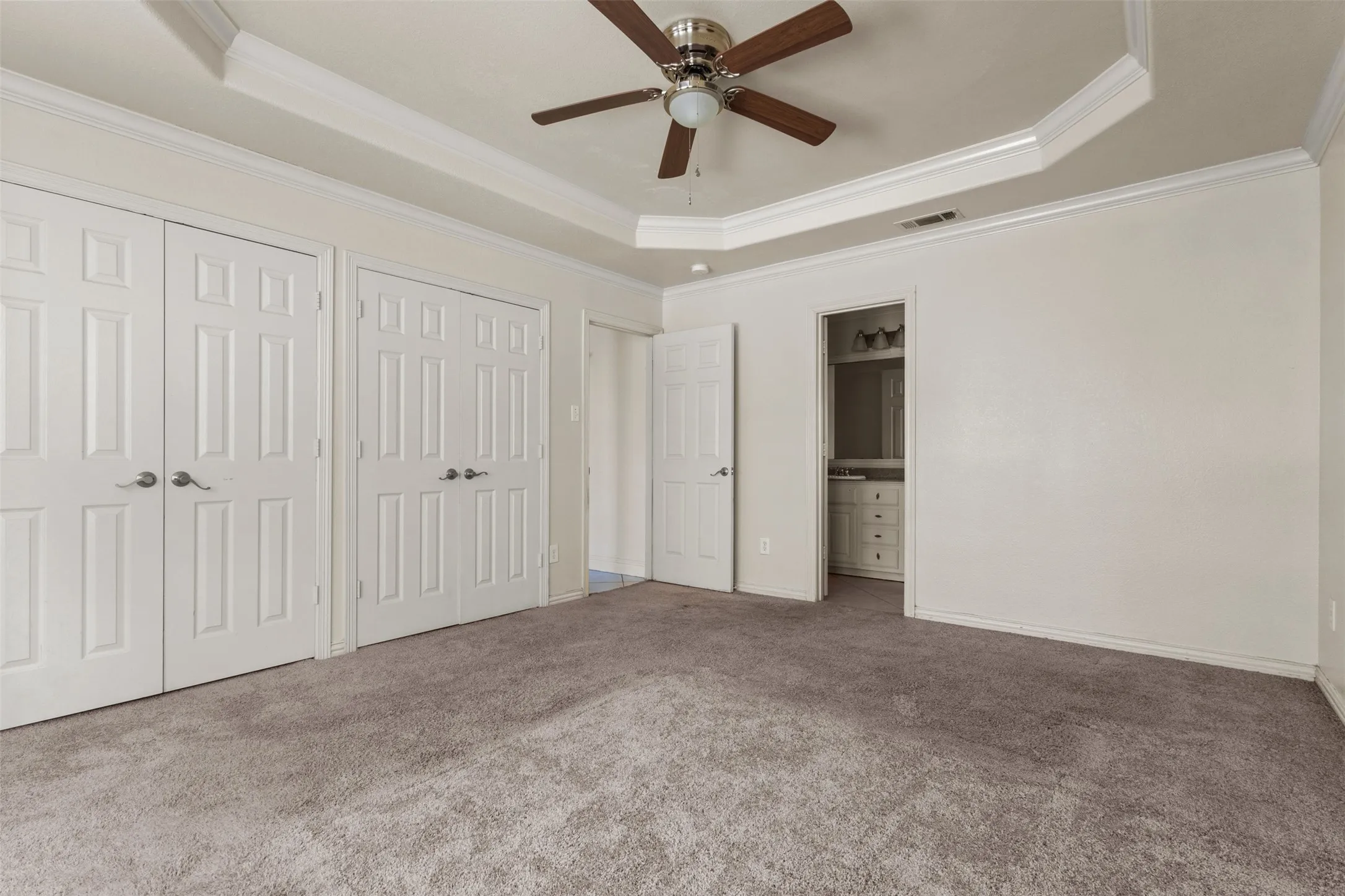 Unfurnished bedroom with a tray ceiling, multiple closets, ensuite bath, carpet, and crown molding