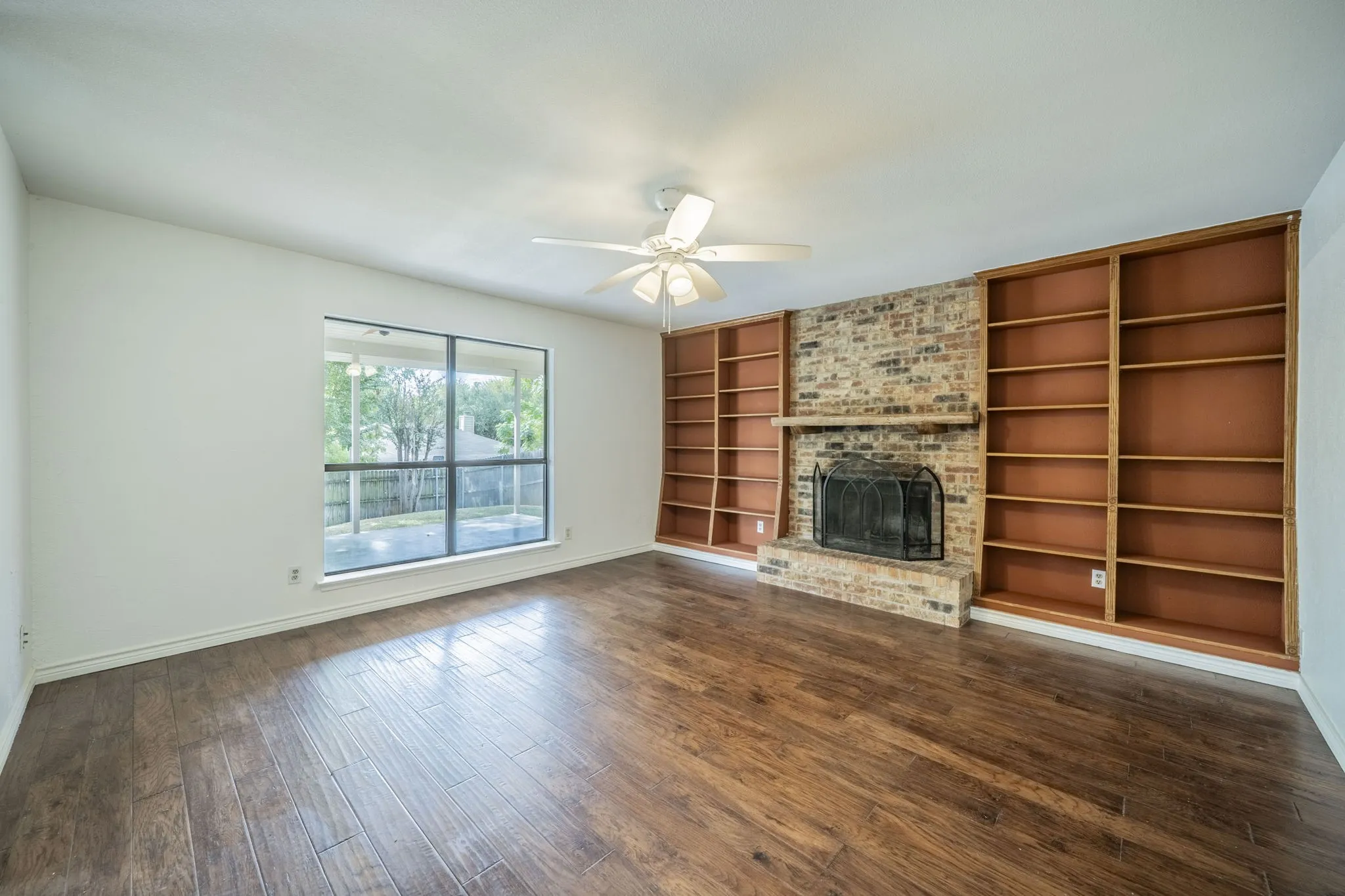 A great living area is at the back of the house and features built in shelves, a fireplace, dark wood-style flooring, and a ceiling fan