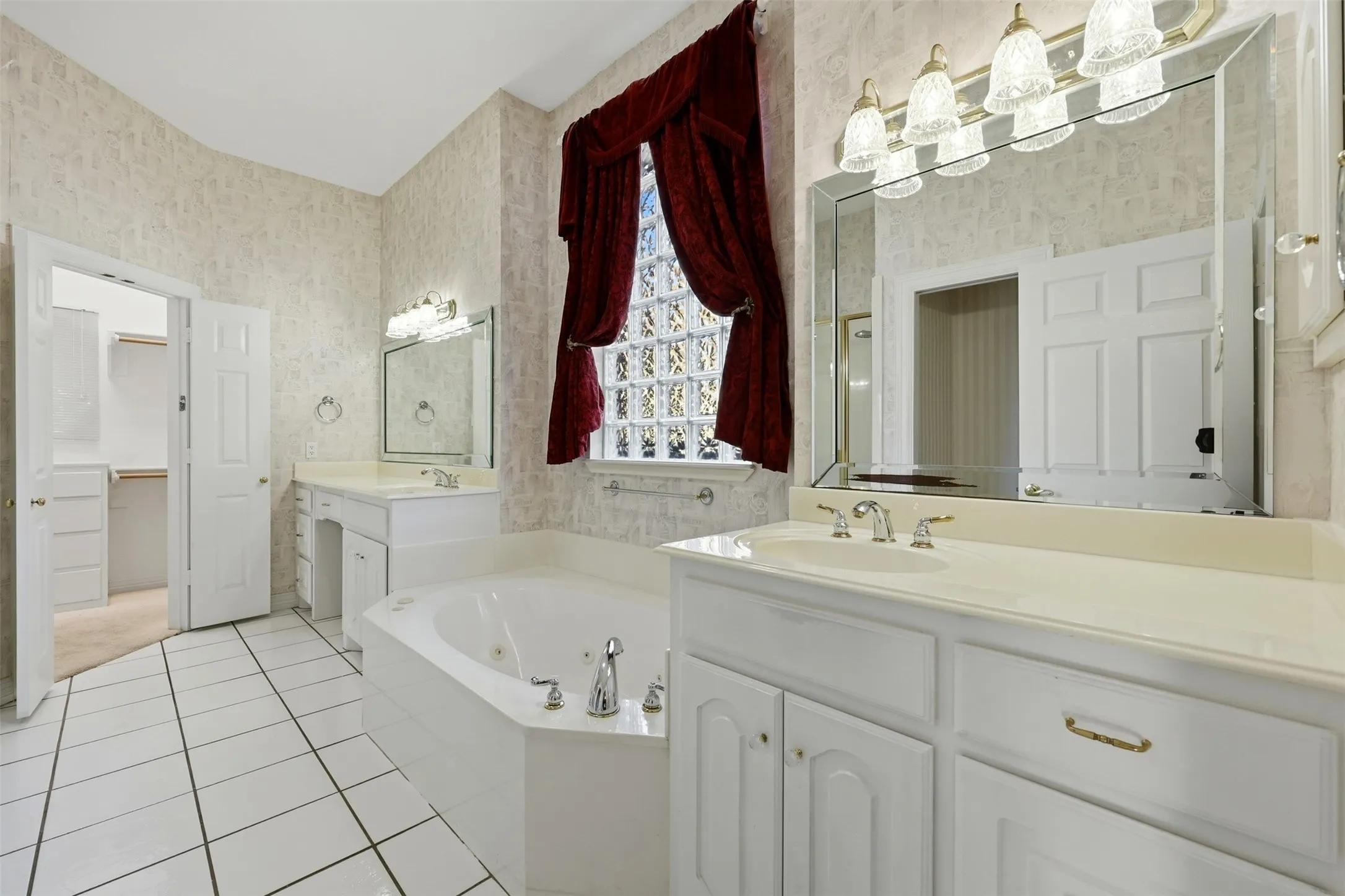 Primary bathroom with wallpapered walls, a jetted tub, light tile patterned floors, two vanities, and a spacious closet
