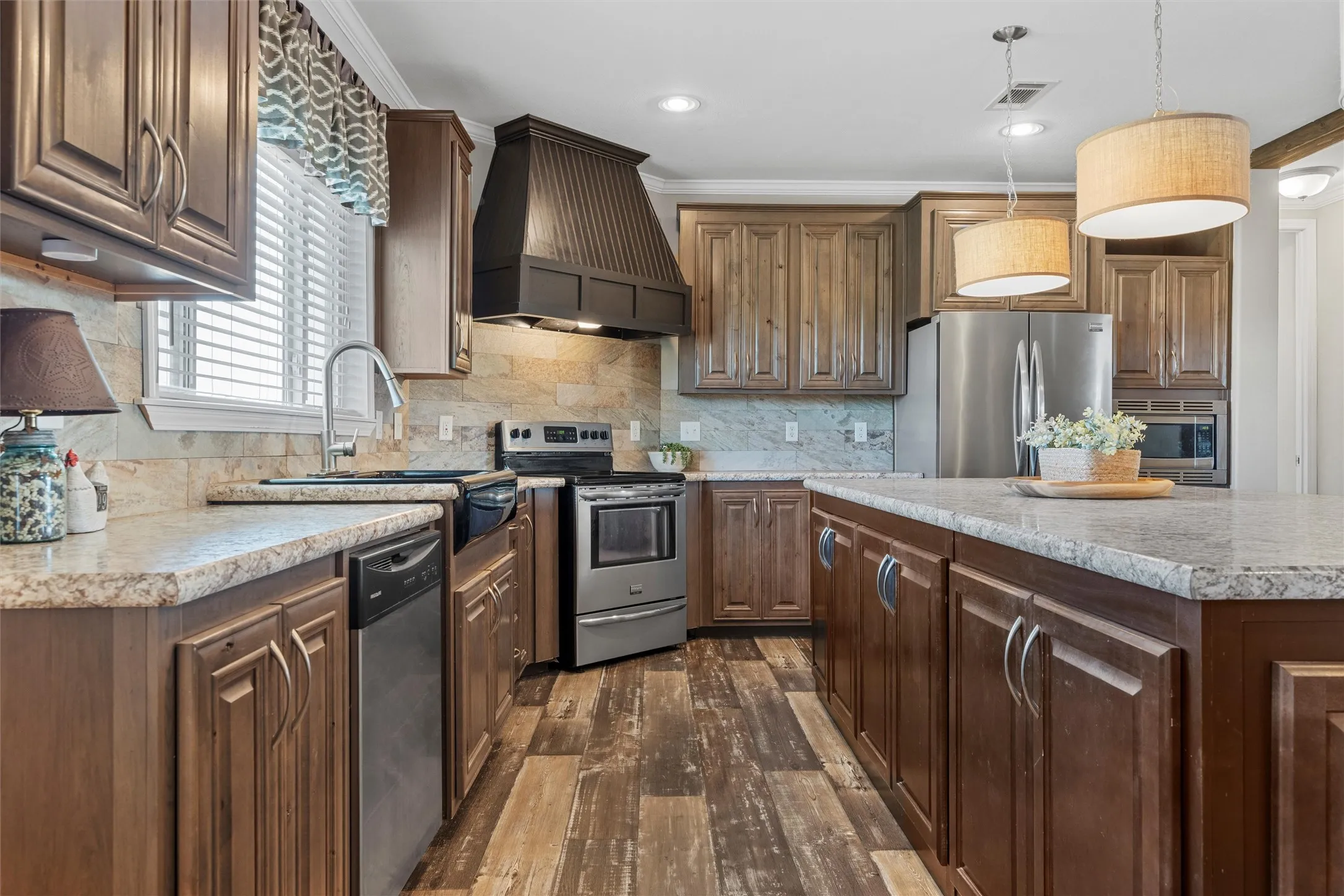 Kitchen featuring custom range hood, appliances with stainless steel finishes, backsplash, dark wood-style flooring, and hanging light fixtures