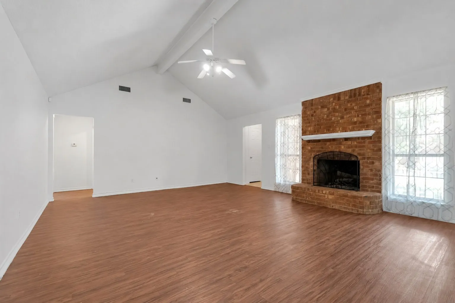 Spacious living area featuring a vaulted ceiling with a ceiling fan, brick fireplace with mantel, and two large windows