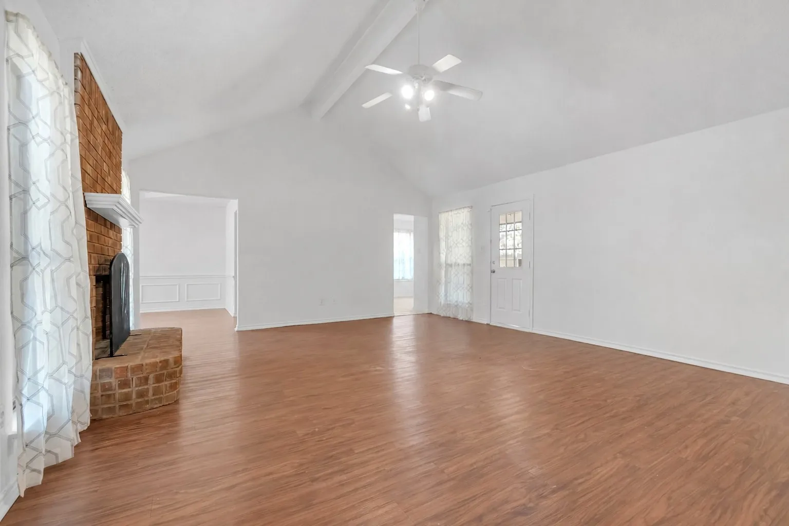Spacious room showcasing gleaming wood-finish flooring throughout, vaulted ceiling with ceiling fan, brick fireplace with mantel, and abundant natural light from multiple windows and glass doors