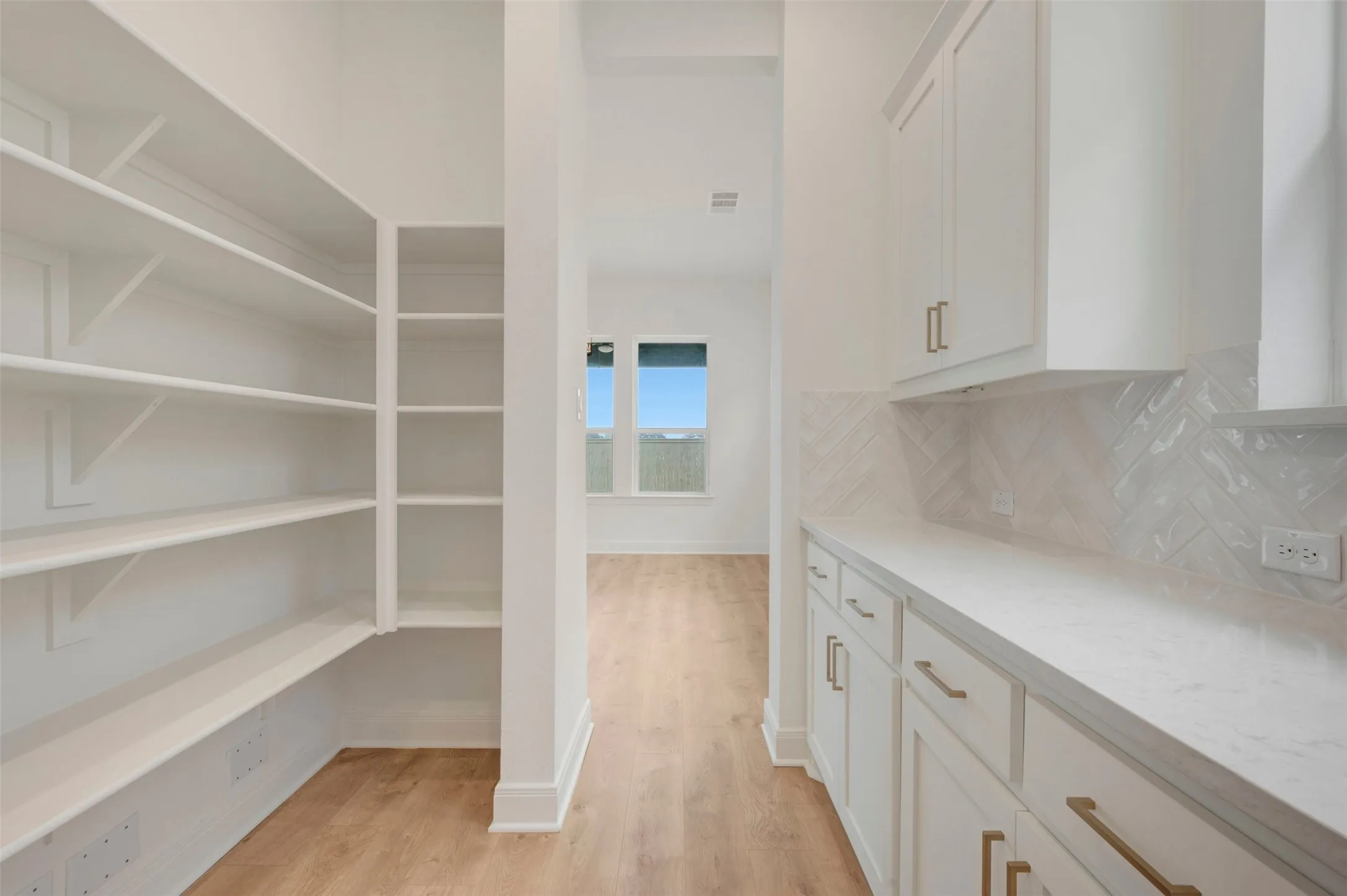 Kitchen pantry featuring extensive built-in shelving, white shaker-style cabinetry with gold-tone hardware, white countertops, and a white herringbone tile backsplash