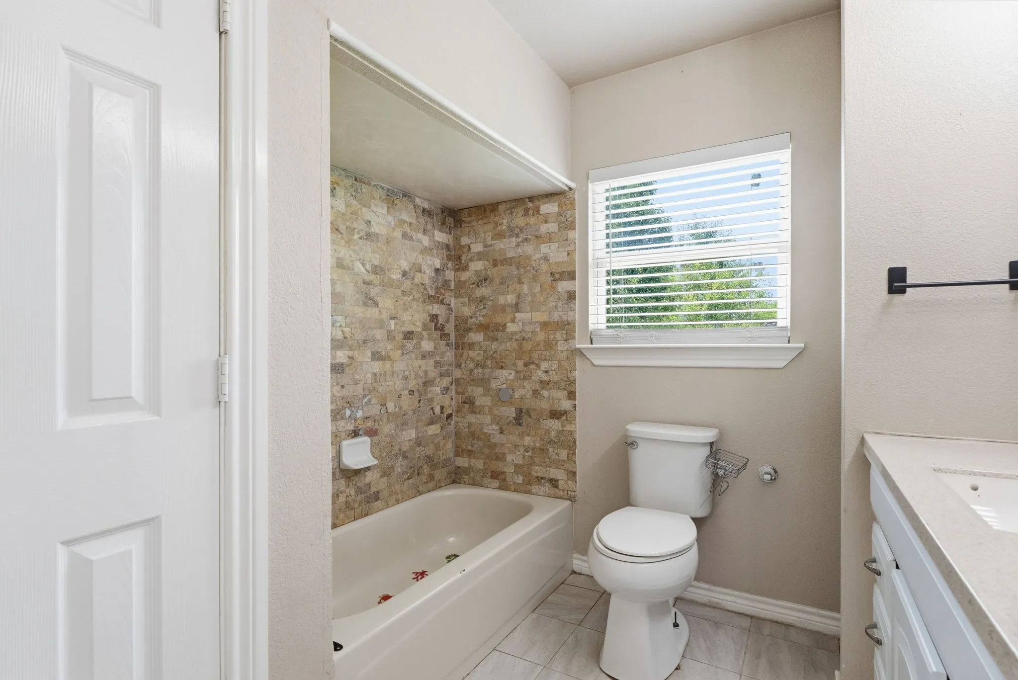 Bathroom featuring a built-in tub with a tiled surround, a toilet, and a window with blinds