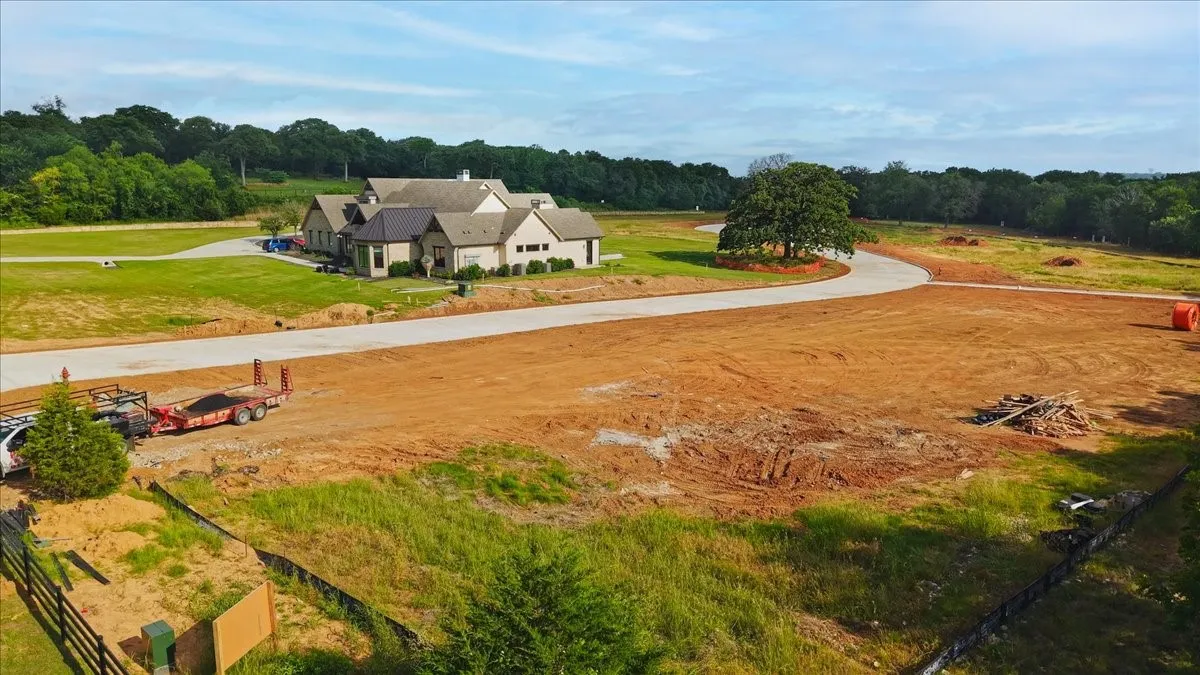 Expansive cleared lot featuring partial fencing, surrounded by established greenery and an asphalt-paved road