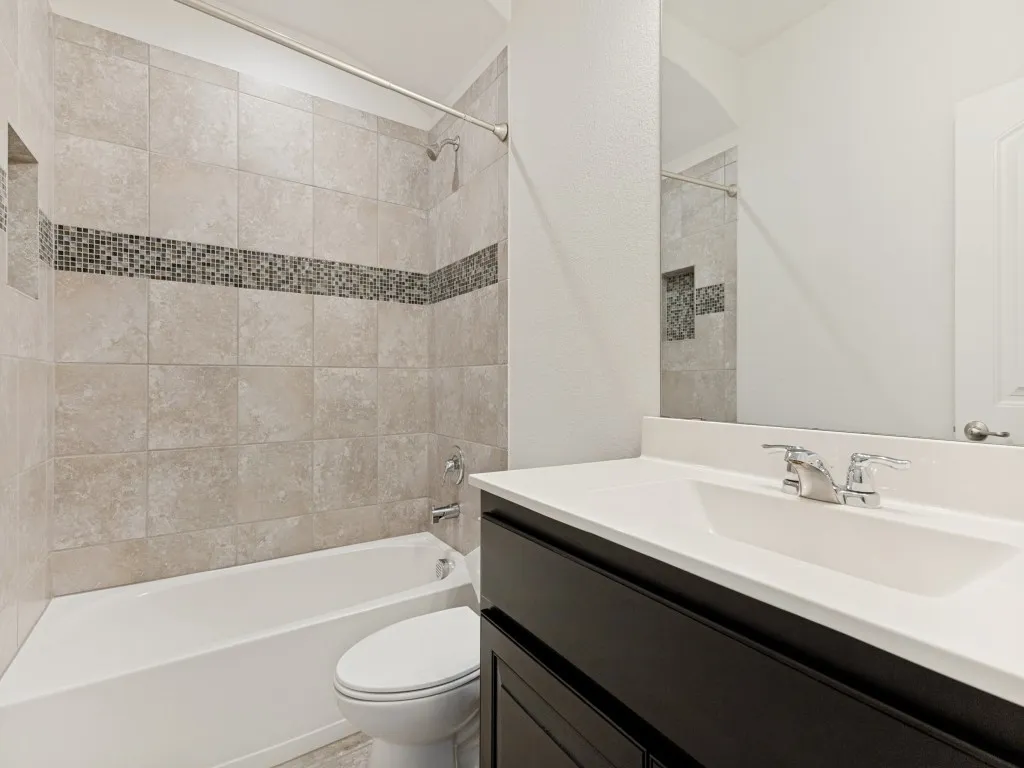 Bathroom featuring a tub/shower combination with beige tile surround and mosaic accent band, a built-in shower niche, dark wood-finish vanity with an integrated white sink and countertop, and a large wall-mounted mirror