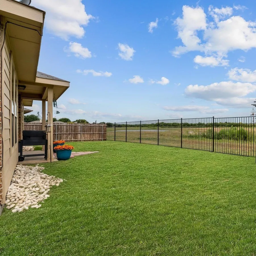 Grassed backyard featuring a black metal fence, a covered patio, and a stone-lined garden bed
