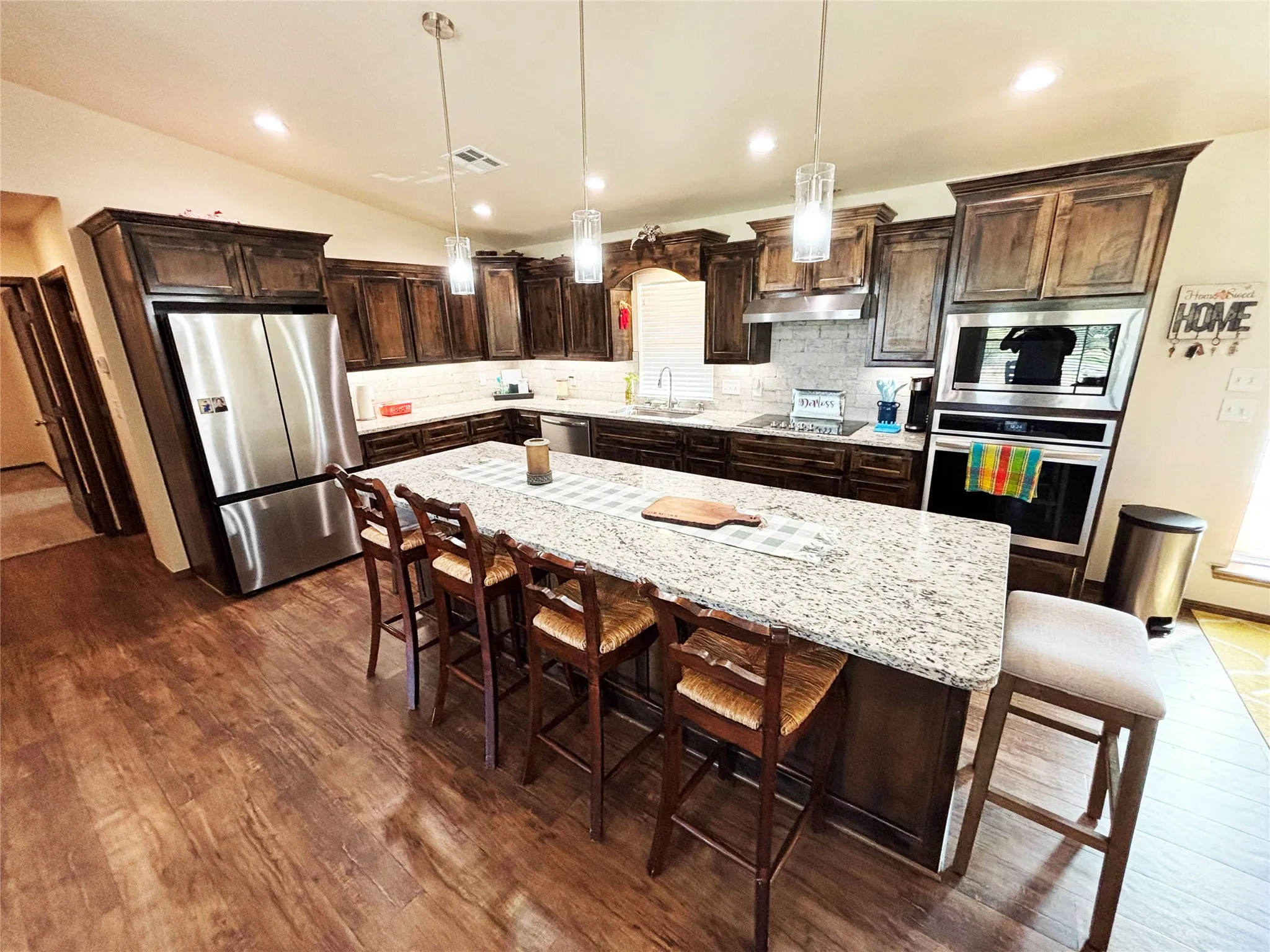Kitchen with dark brown cabinets, decorative light fixtures, stainless steel appliances, light stone countertops, and a breakfast bar area