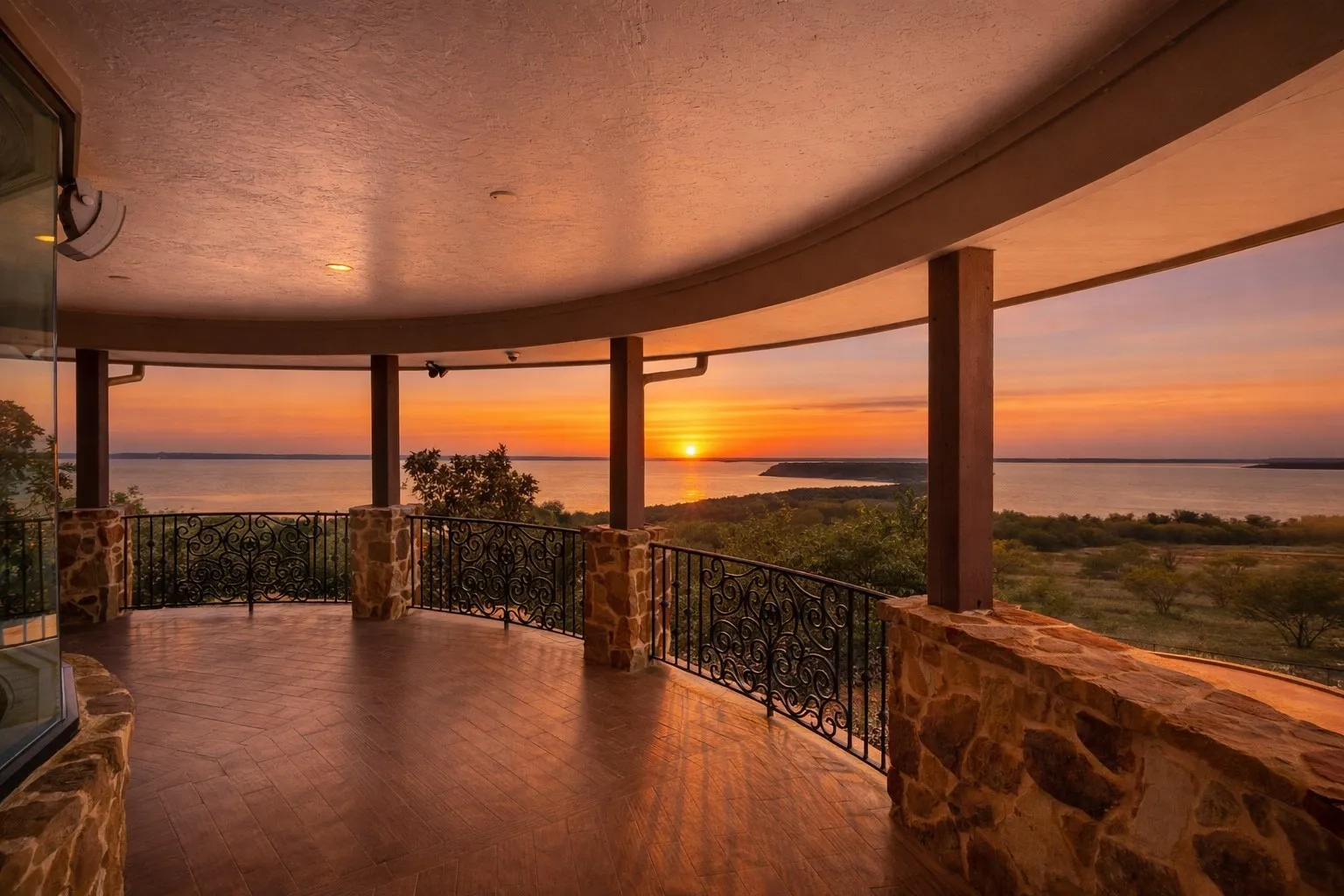 Curved covered balcony featuring a wood-finish flooring, stone pillars, and ornate wrought iron railings overlooking a prominent body of water and distant land formations