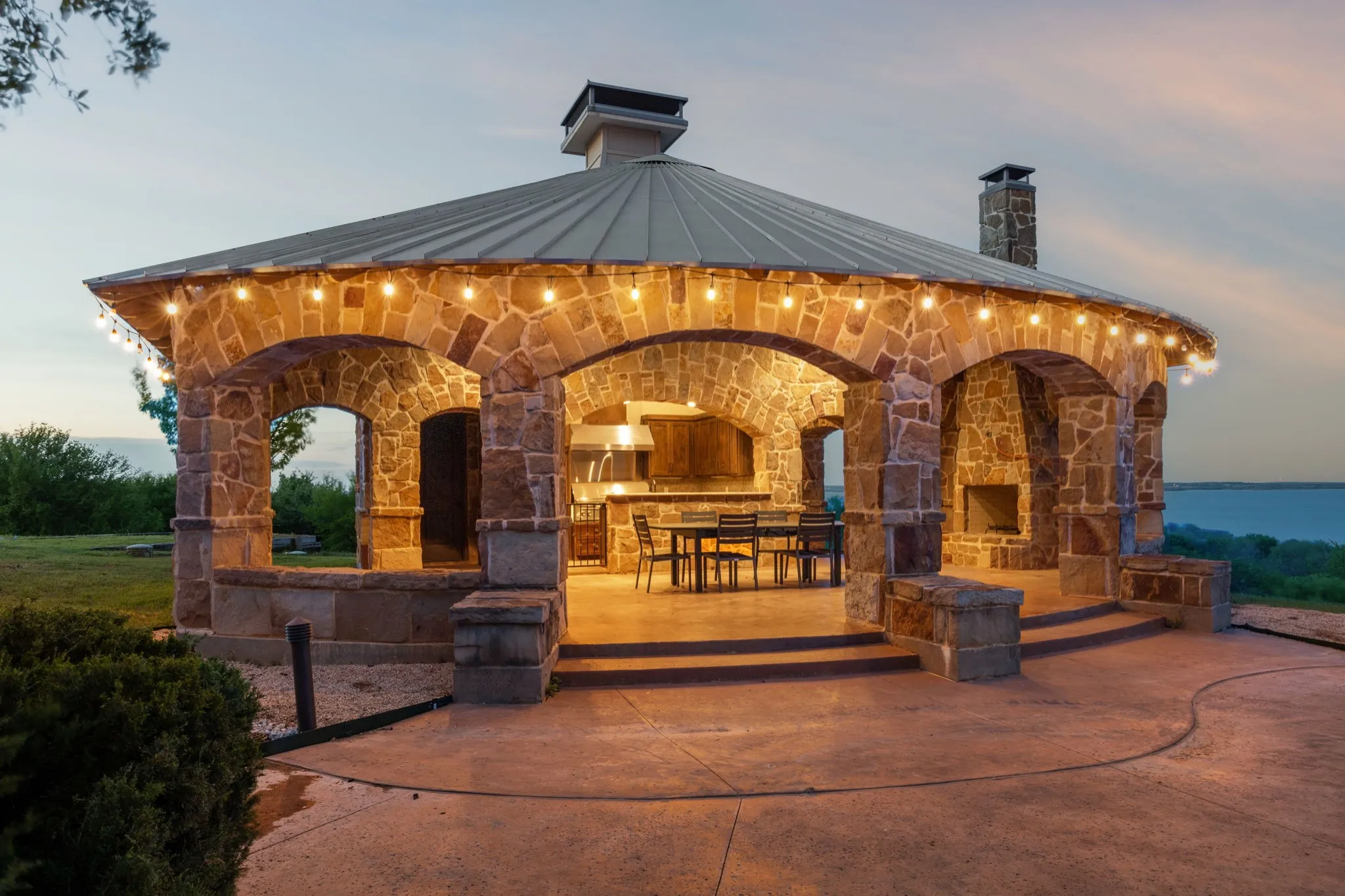 Stone-clad outdoor pavilion featuring a metal roof, arched openings, built-in fireplace, and integrated kitchen area