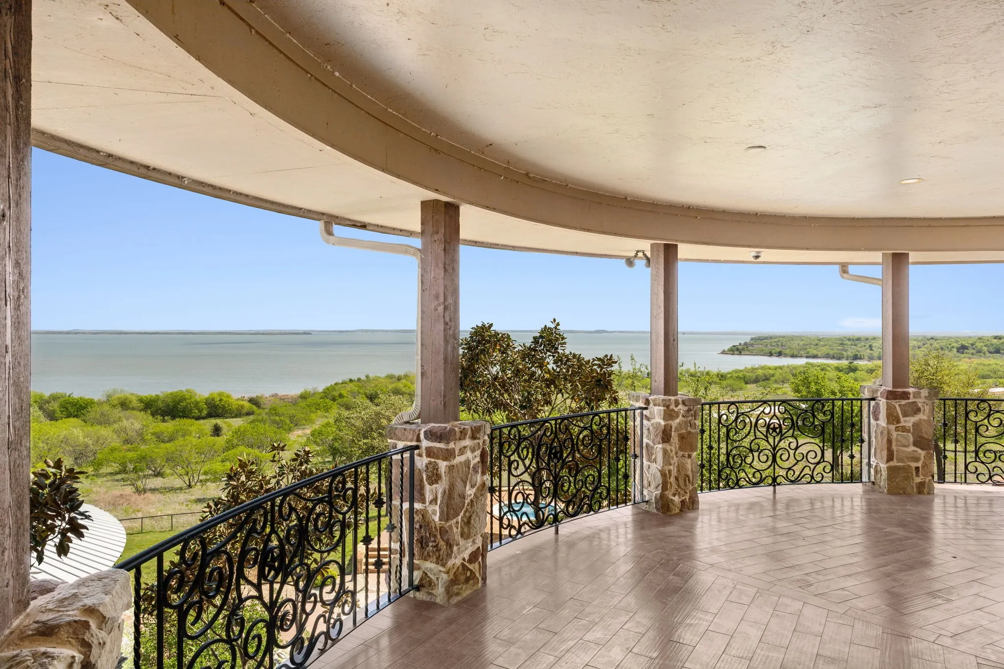 Curved covered patio featuring stone columns, decorative wrought iron railing, and herringbone-patterned tile flooring