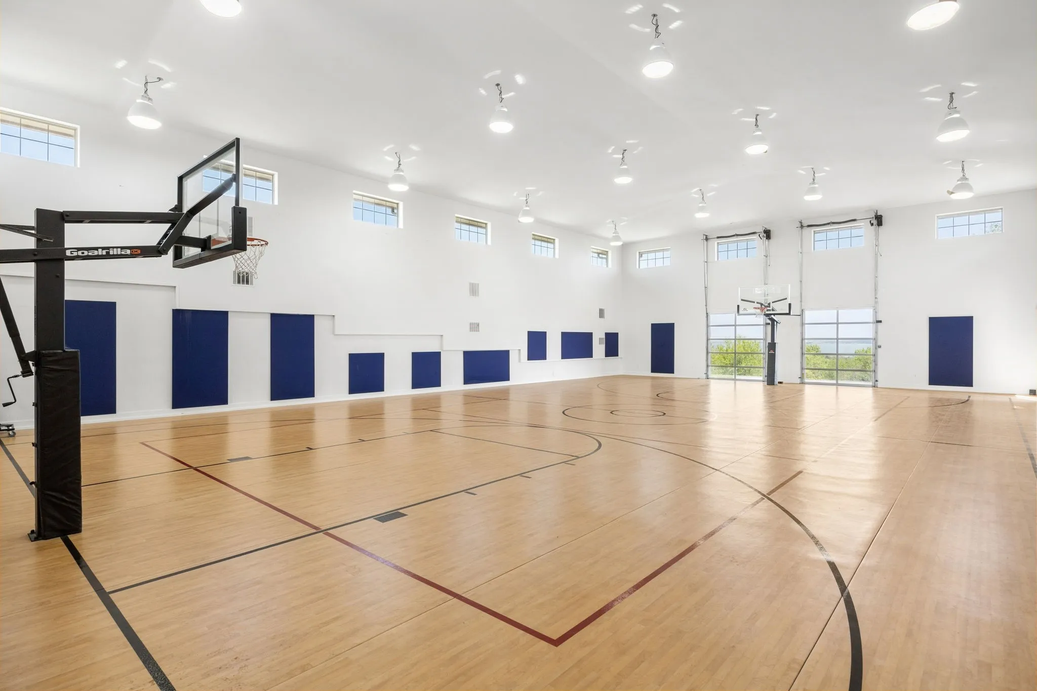 Indoor basketball court featuring wood-finish flooring, padded wall panels, multiple transom windows, and overhead task lighting