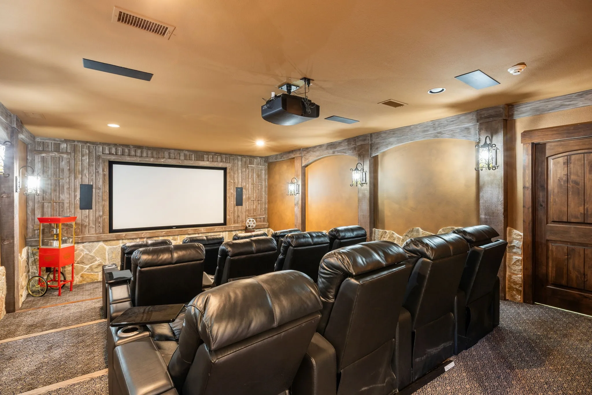 Dedicated media room featuring a large projection screen, tiered seating, wood-paneled walls, stone accents, and wall sconce lighting