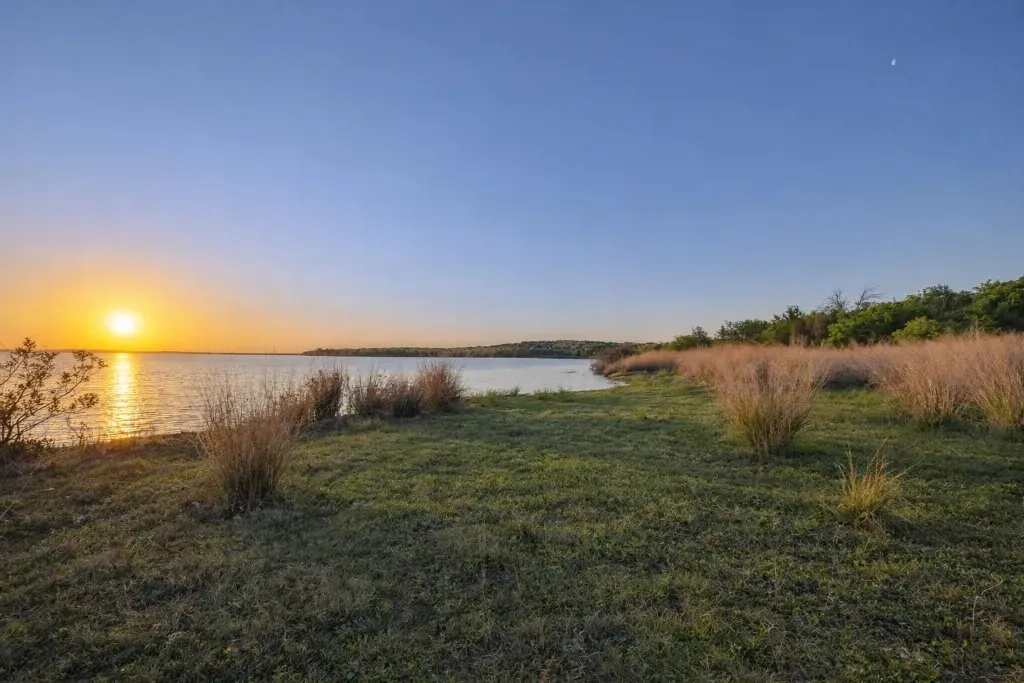 Expansive waterfront property featuring a calm body of water, a natural shoreline with green grass, and mature foliage bordering the horizon