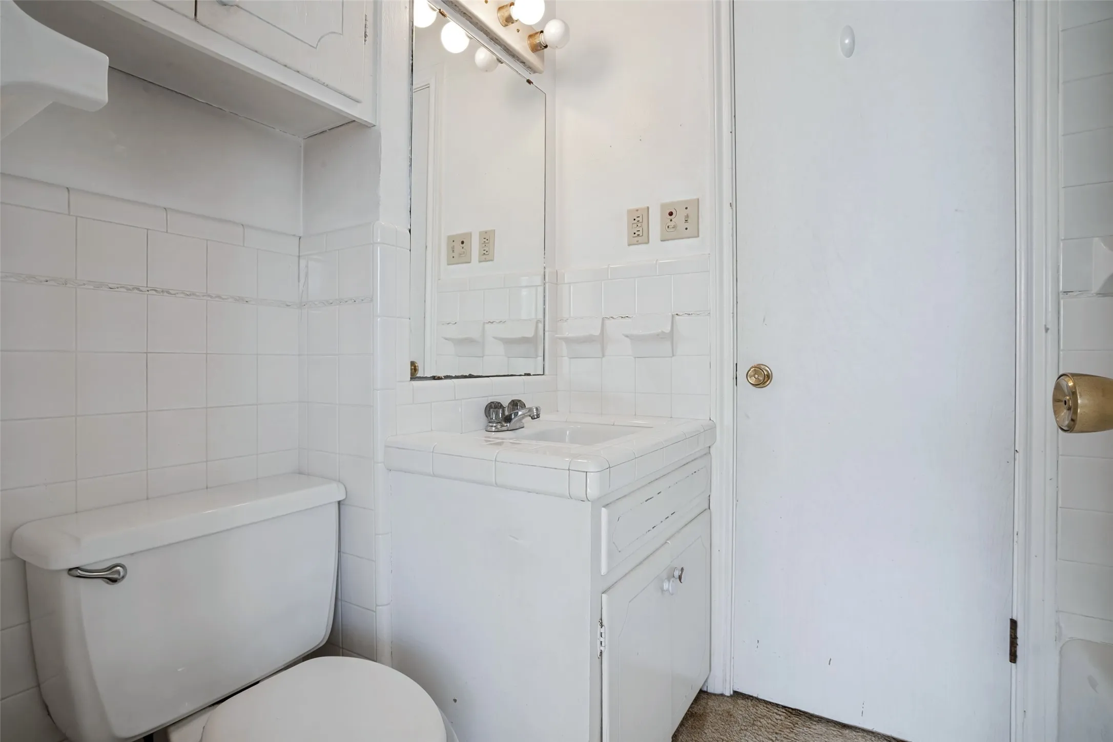 White tile walls, built-in medicine cabinet, vanity with integrated sink, overhead vanity lighting, white door with brass-finish hardware