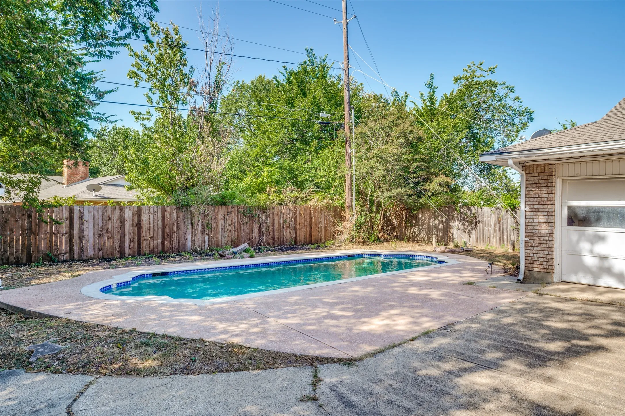 In-ground swimming pool with surrounding concrete decking, wood fence perimeter, and adjacent brick exterior with white garage door
