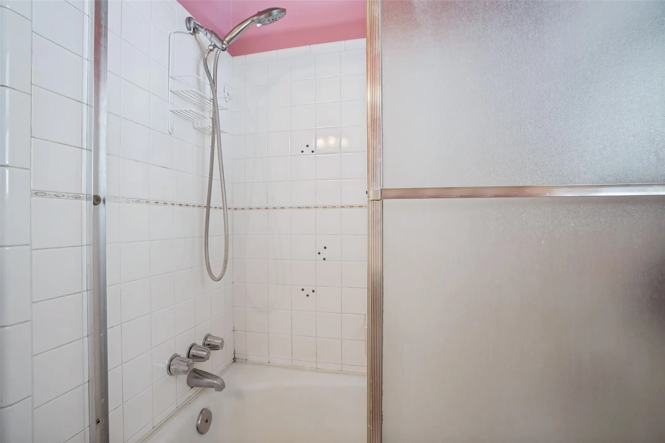 Bathtub with white tile surround, a handheld shower fixture, and sliding frosted glass enclosure