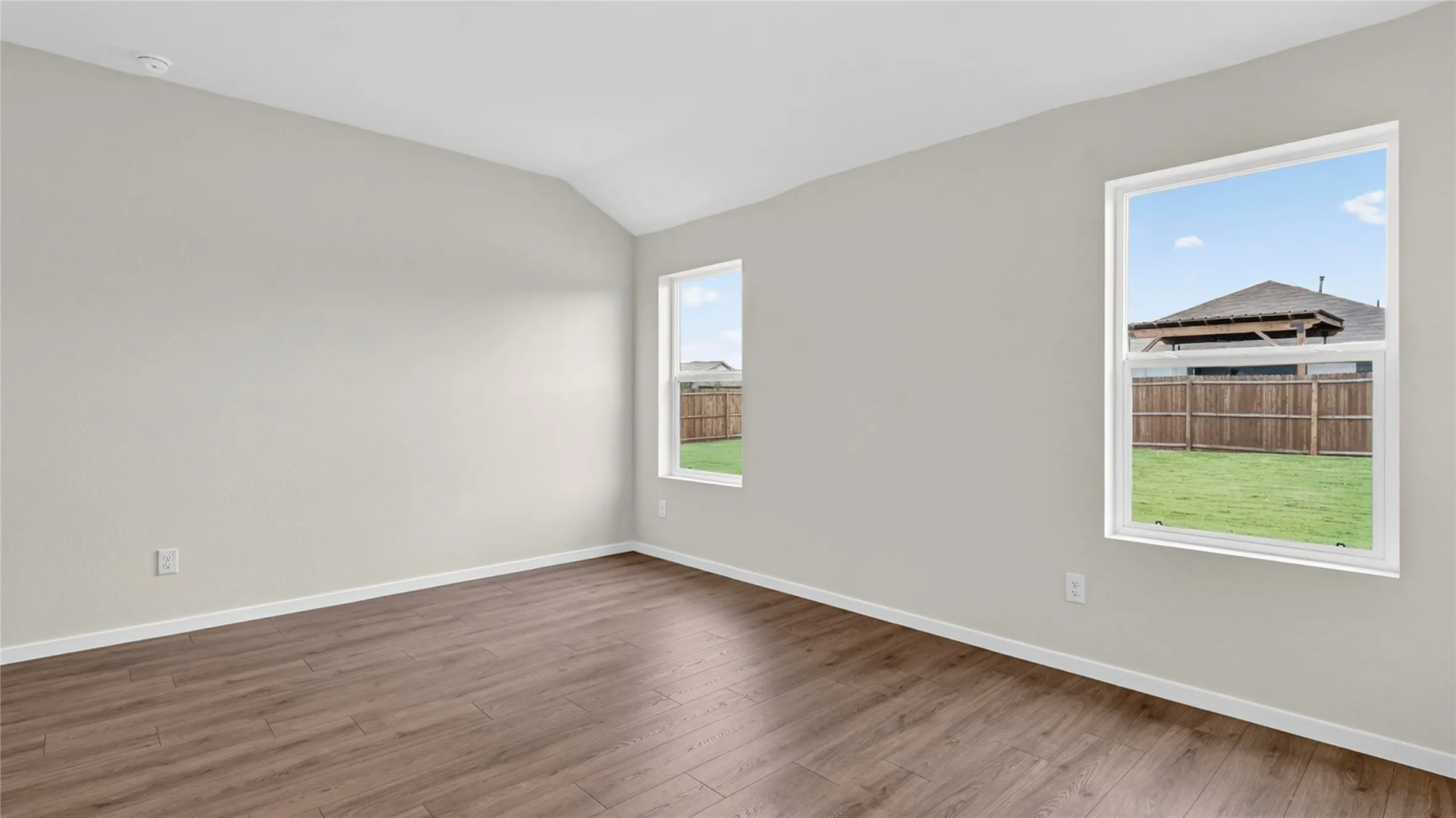 Vaulted ceiling room featuring wood-finish flooring, light neutral wall paint, and two windows providing natural light