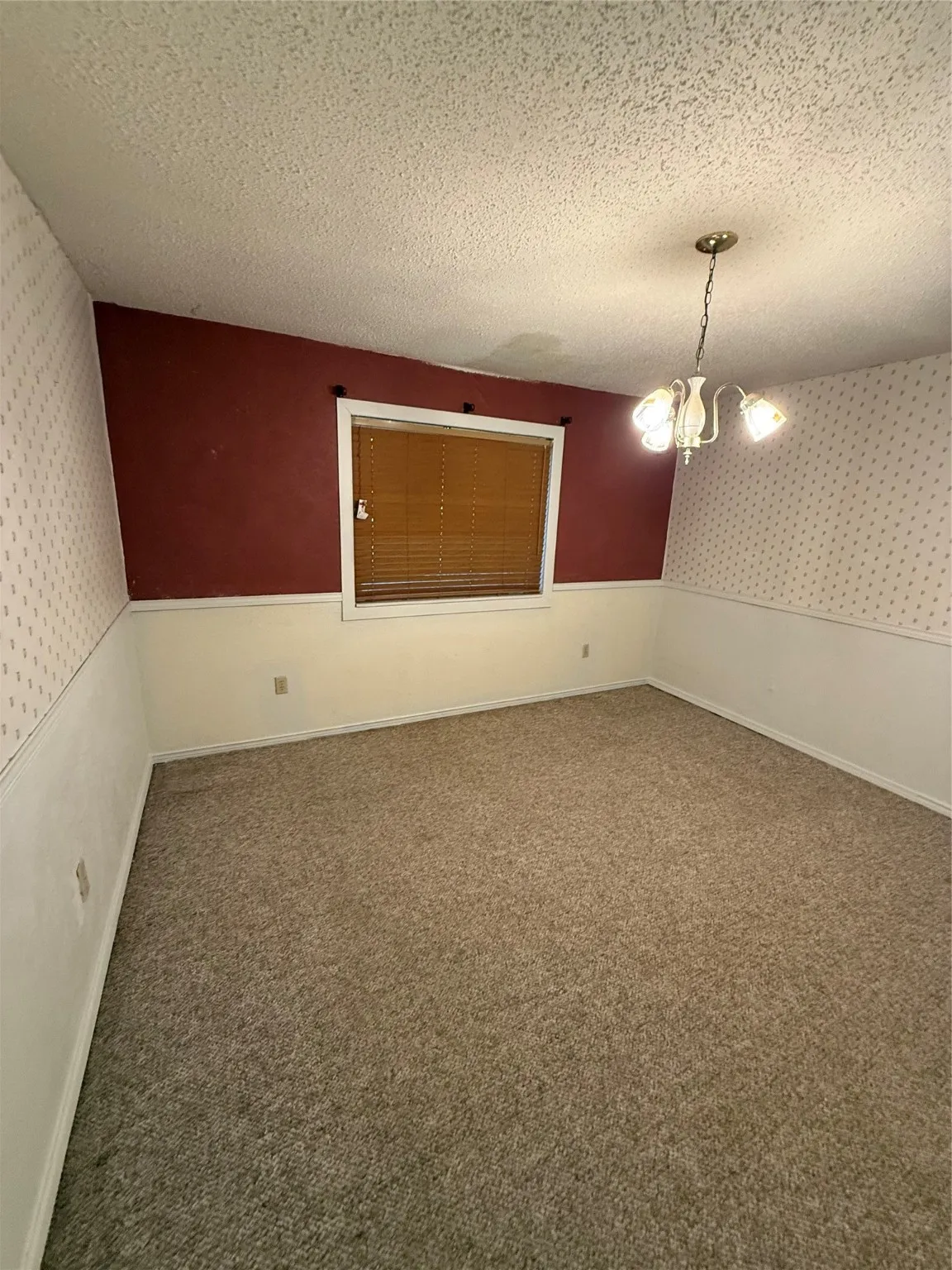 Carpeted spare room with wallpapered walls, a textured ceiling, a chandelier, and a wainscoted wall
