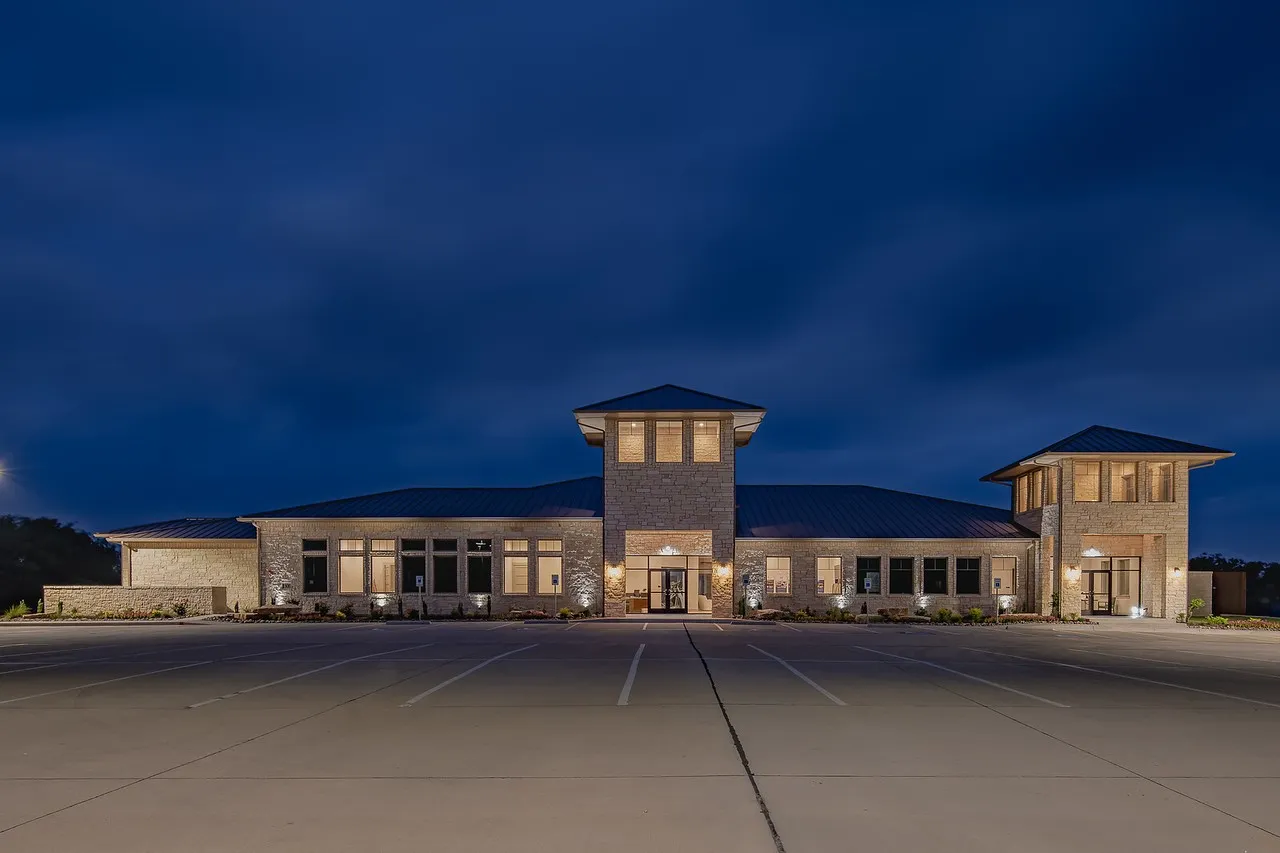 Contemporary stone facade building with two prominent towers, dark metal roofing, abundant window installations, glass entry doors, and extensive paved parking