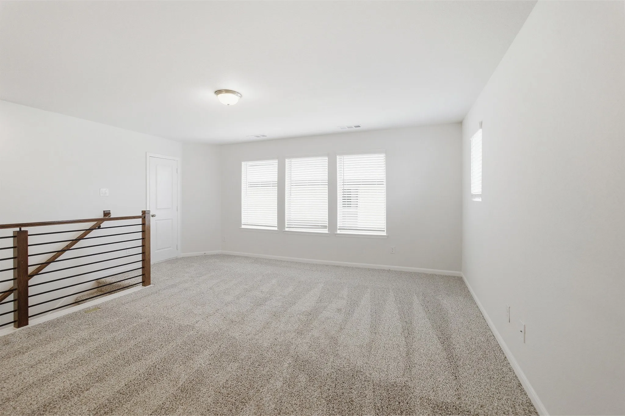 Expansive carpeted loft space featuring multiple windows with blinds, a sleek horizontal railing, and a flush mount ceiling light fixture