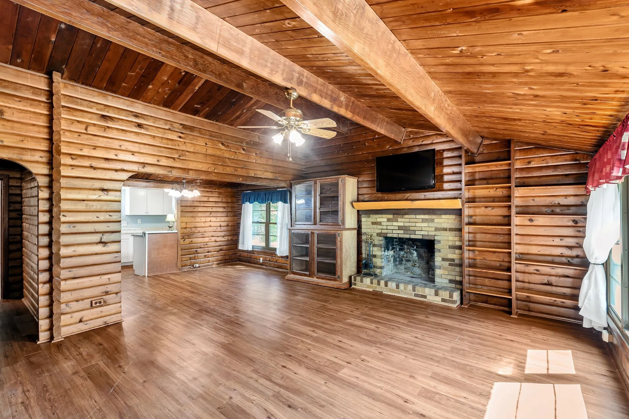 Living room featuring a brick fireplace, log walls, wood finished floors, wooden ceiling, and ceiling fan