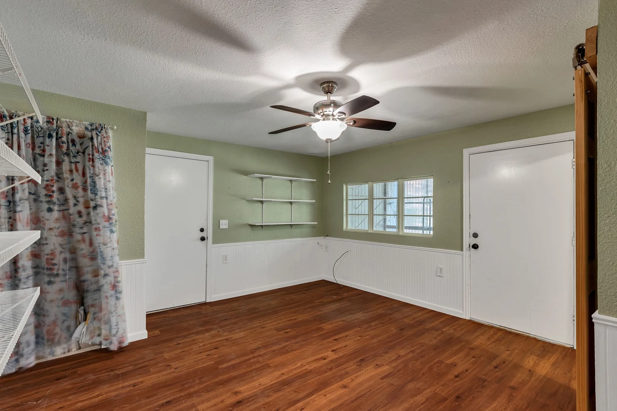 Bedroom with attached full bathroom featuring dark wood-style flooring, a wainscoted wall, ceiling fan, and a barn door