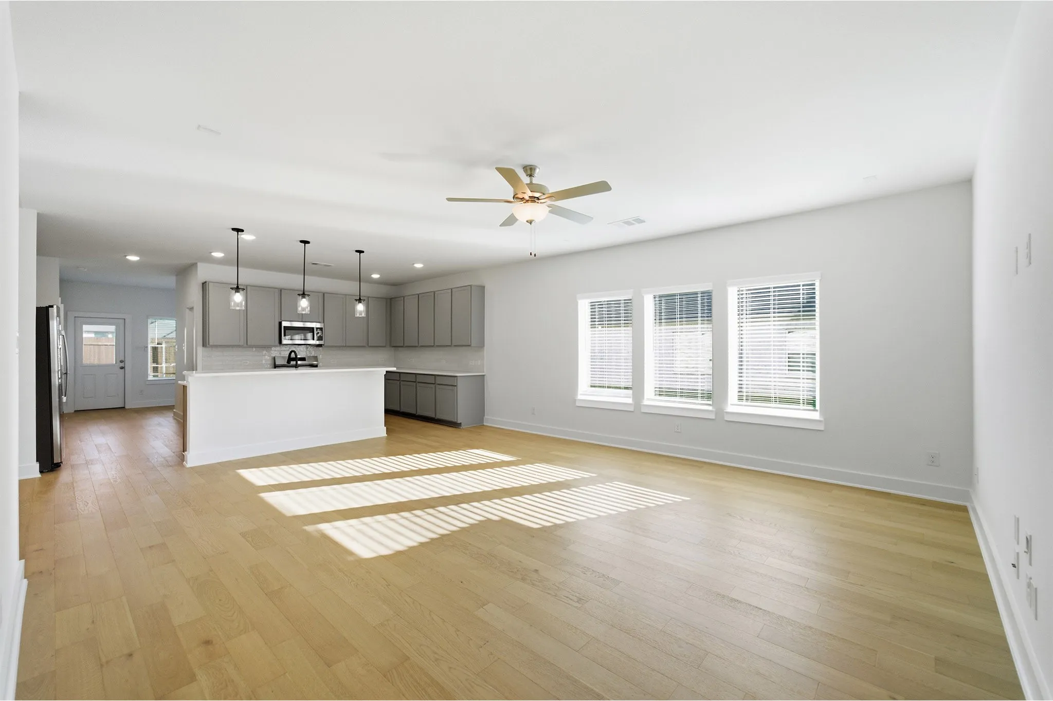 Open-concept living area featuring wood-finish flooring, a kitchen island with pendant lighting, gray cabinetry, and three large windows