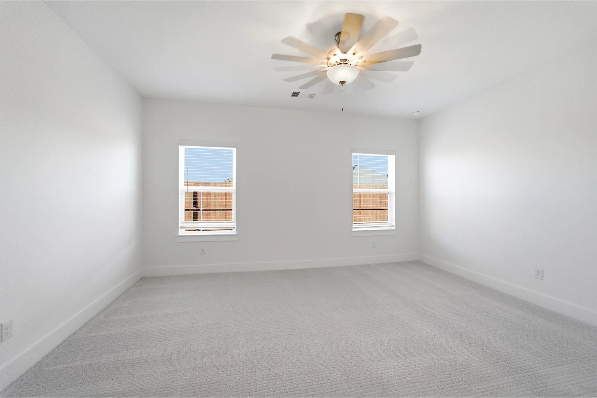 Spacious room featuring light gray carpeting, crisp white walls, two windows with white blinds, a white ceiling fan with integrated lighting, and white baseboards